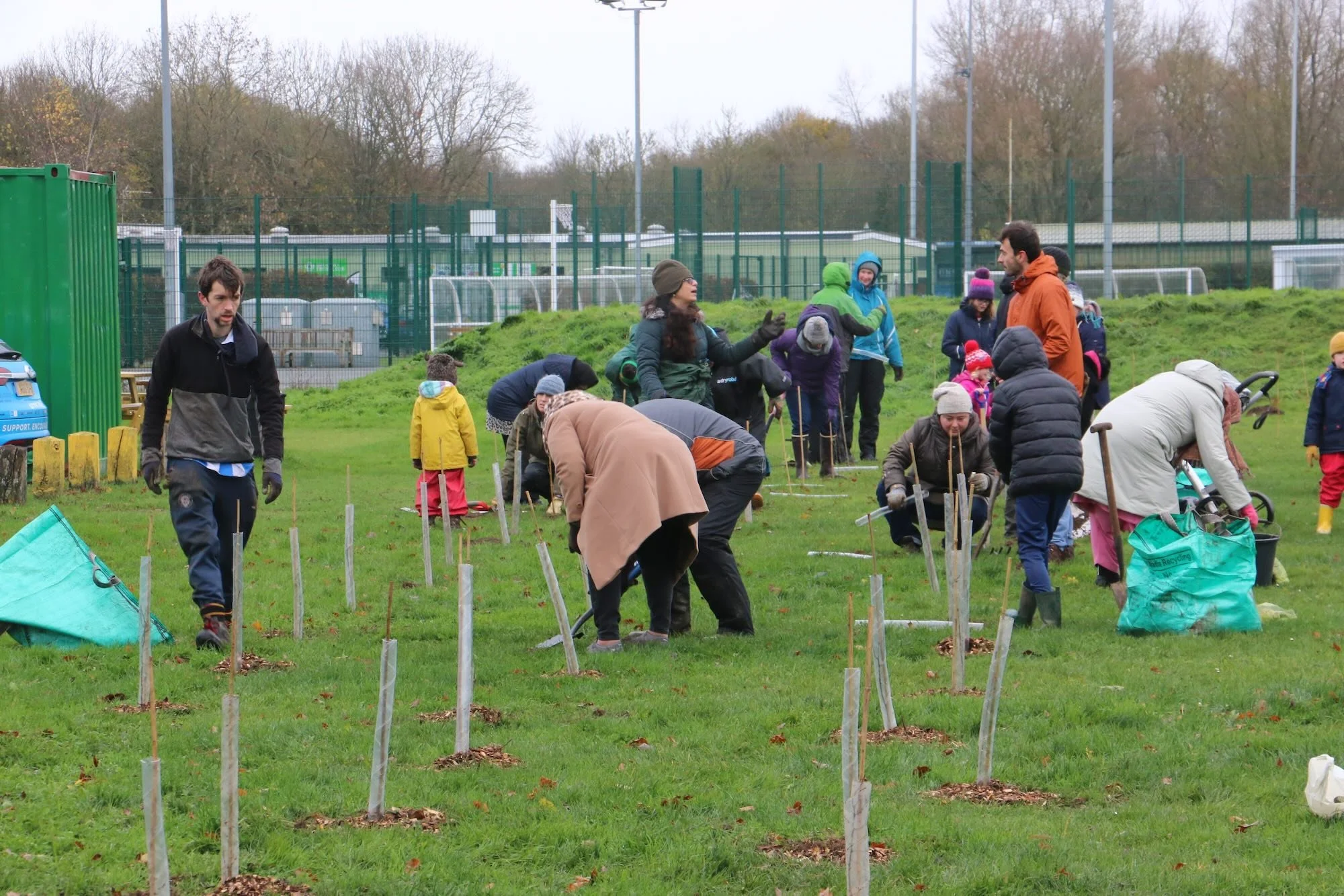 Group of people planting trees on a grassy field on a cloudy day. Some are bending down, others are standing and talking, and children are also present. The background shows sports fields and a green fence.