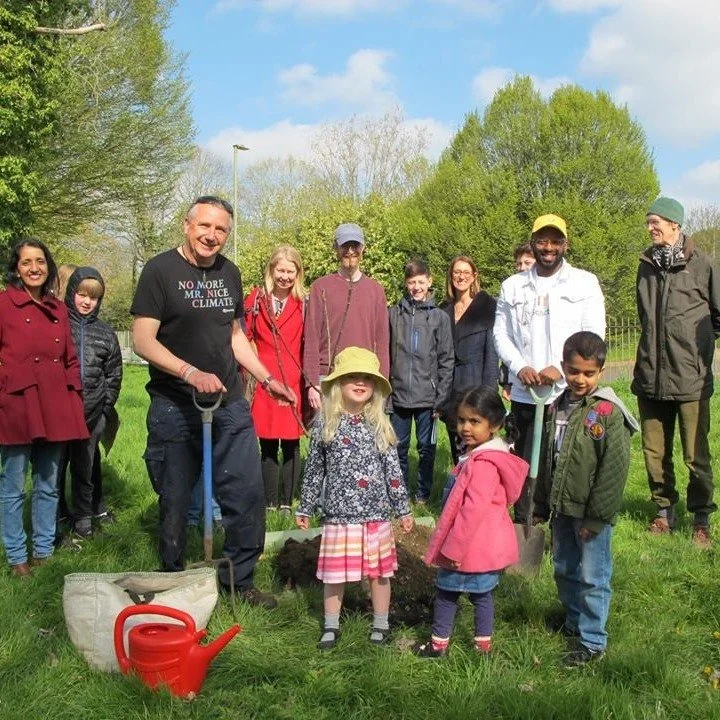 A group of people, including children and adults, gathered outdoors on a grassy area with trees in the background. Some are holding shovels and participating in a planting activity, with a young girl in a sunhat and a little girl in a pink jacket in the foreground. A red watering can is on the ground nearby.