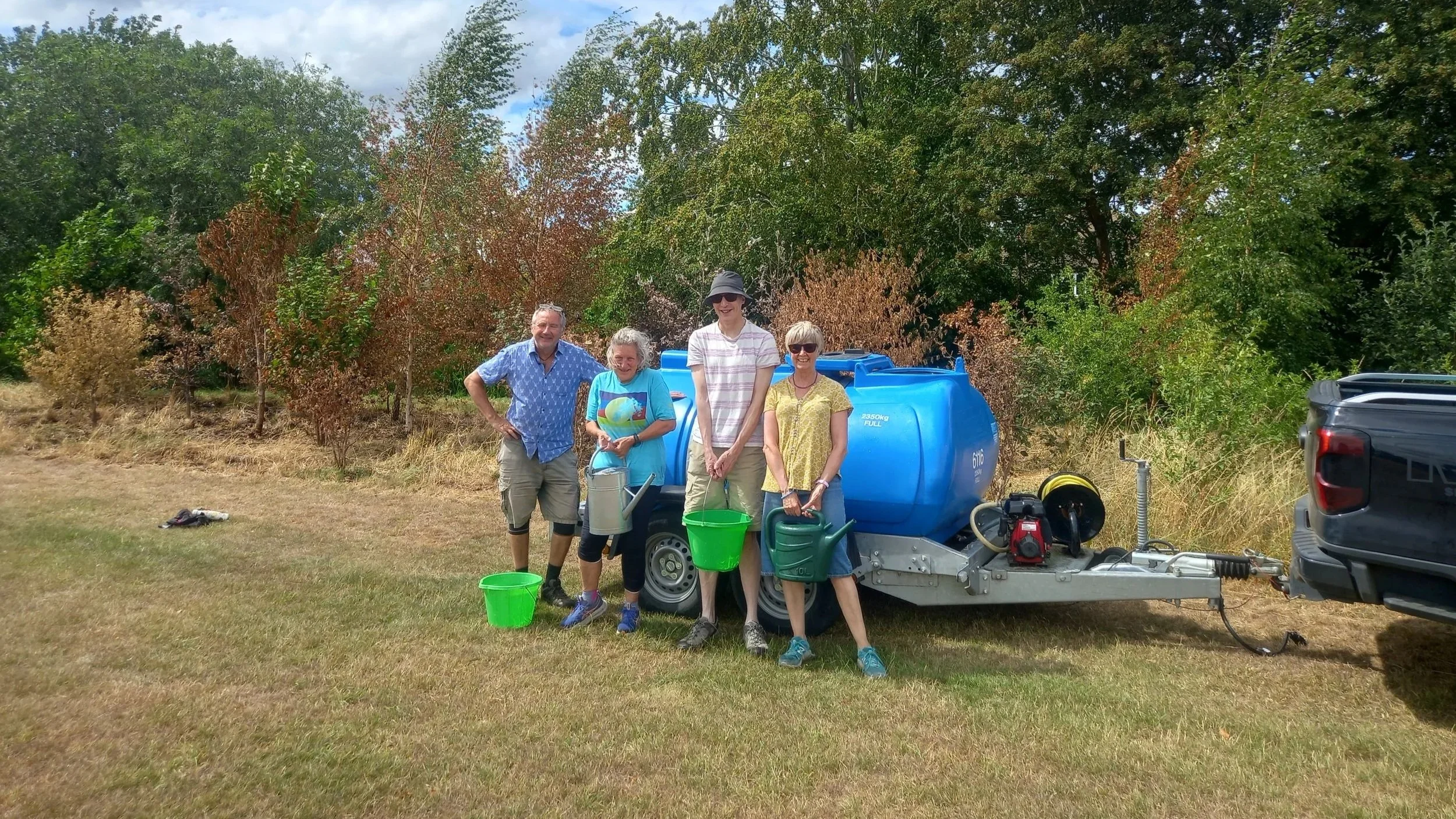 Four volunteers holding buckets and watering cans beside a large blue water tank, with drought-stressed young trees behind them.