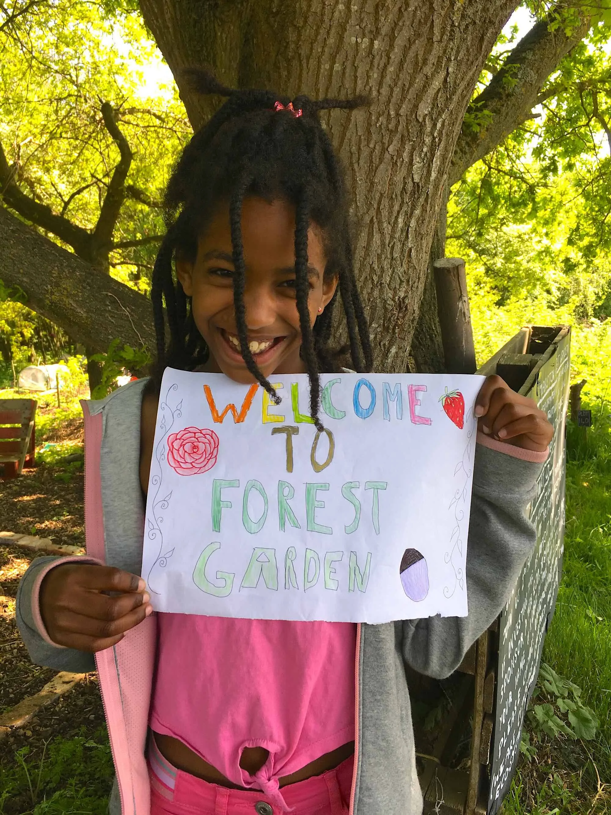 A smiling young girl with dreadlocks, wearing a pink shirt and a light gray jacket, holding a colorful handwritten sign that says 'Welcome to Forest Garden' with illustrations of a flower and an eggplant, standing outdoors next to a large tree with green leaves.
