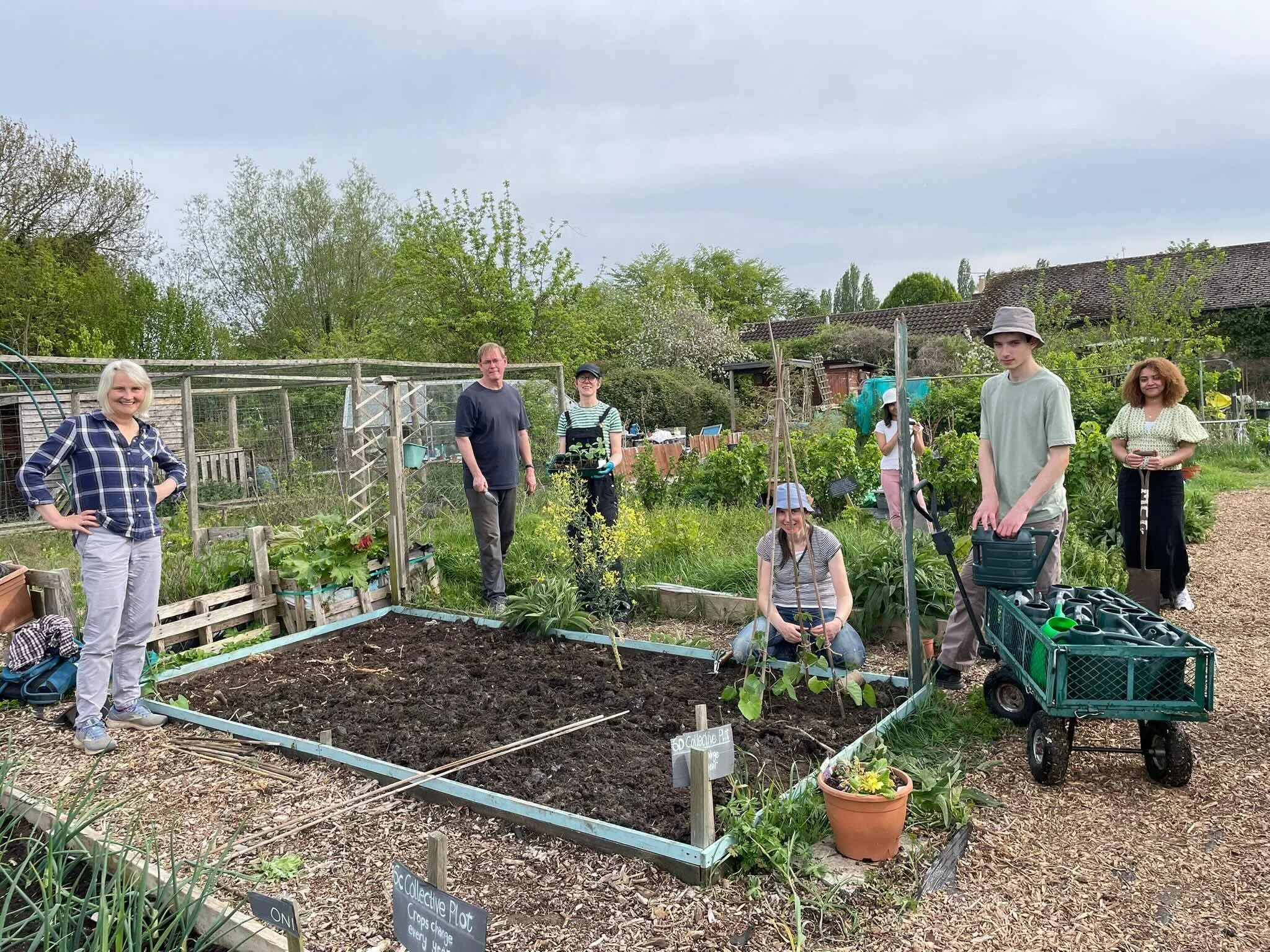 Group of people standing in a community garden with plants and gardening tools, smiling at the camera under cloudy sky.