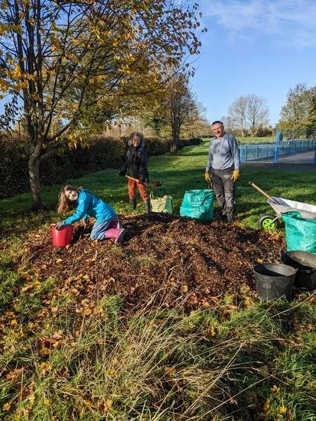 Three people working together outdoors, planting or tending to a garden bed with soil. They are near a tree with autumn leaves and a fence in the background, under a partly cloudy sky.