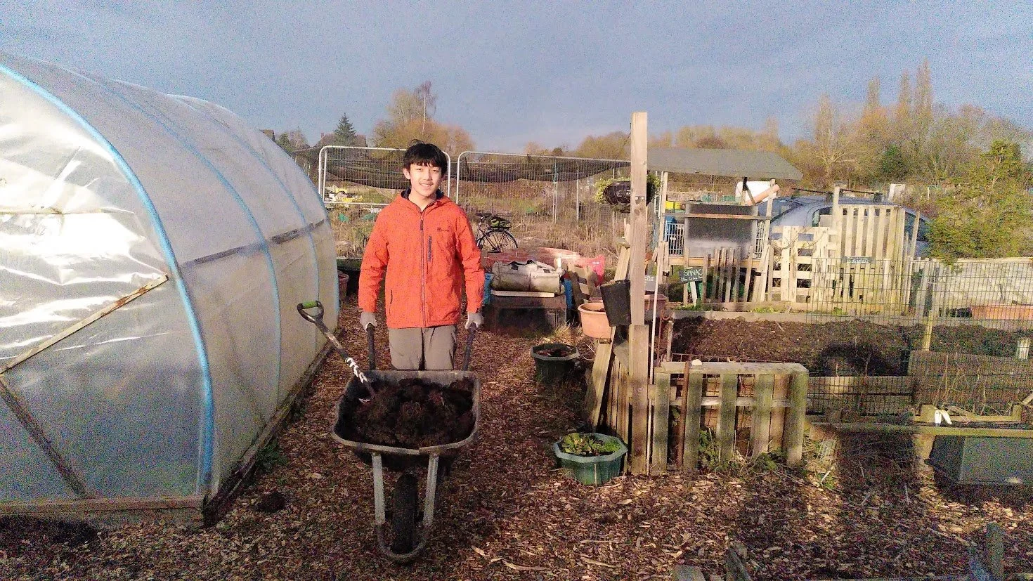 A person in an orange jacket and gray pants stands in a garden, pushing a wheelbarrow filled with dark soil, surrounded by gardening tools and supplies.