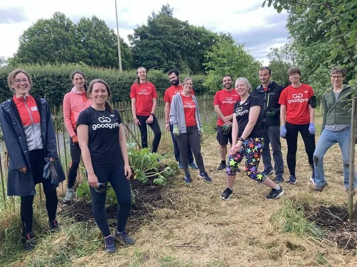 Group of people smiling outdoors during a gardening event, some holding tools, plants, and gloves, with greenery and trees in the background.