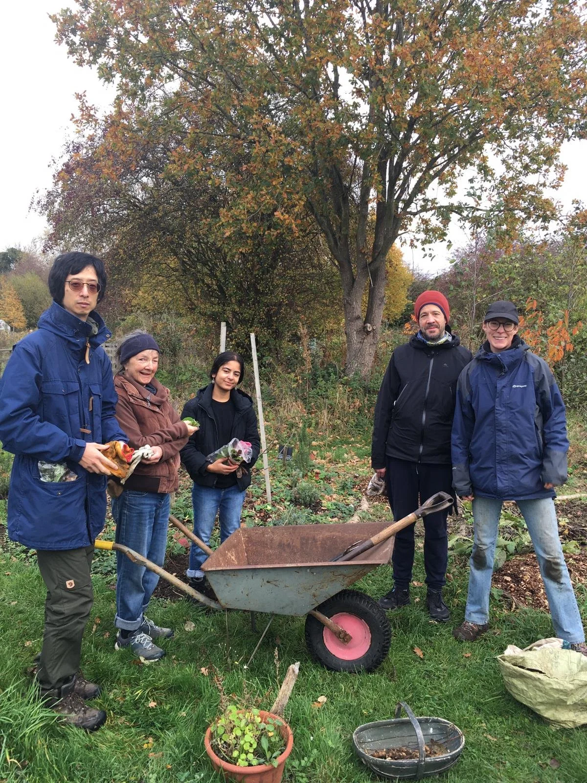 Group of five people gardening outdoors on a cloudy autumn day, with trees displaying fall foliage in the background. They are standing near a wheelbarrow, holding gardening tools and plants, dressed in jackets and hats.