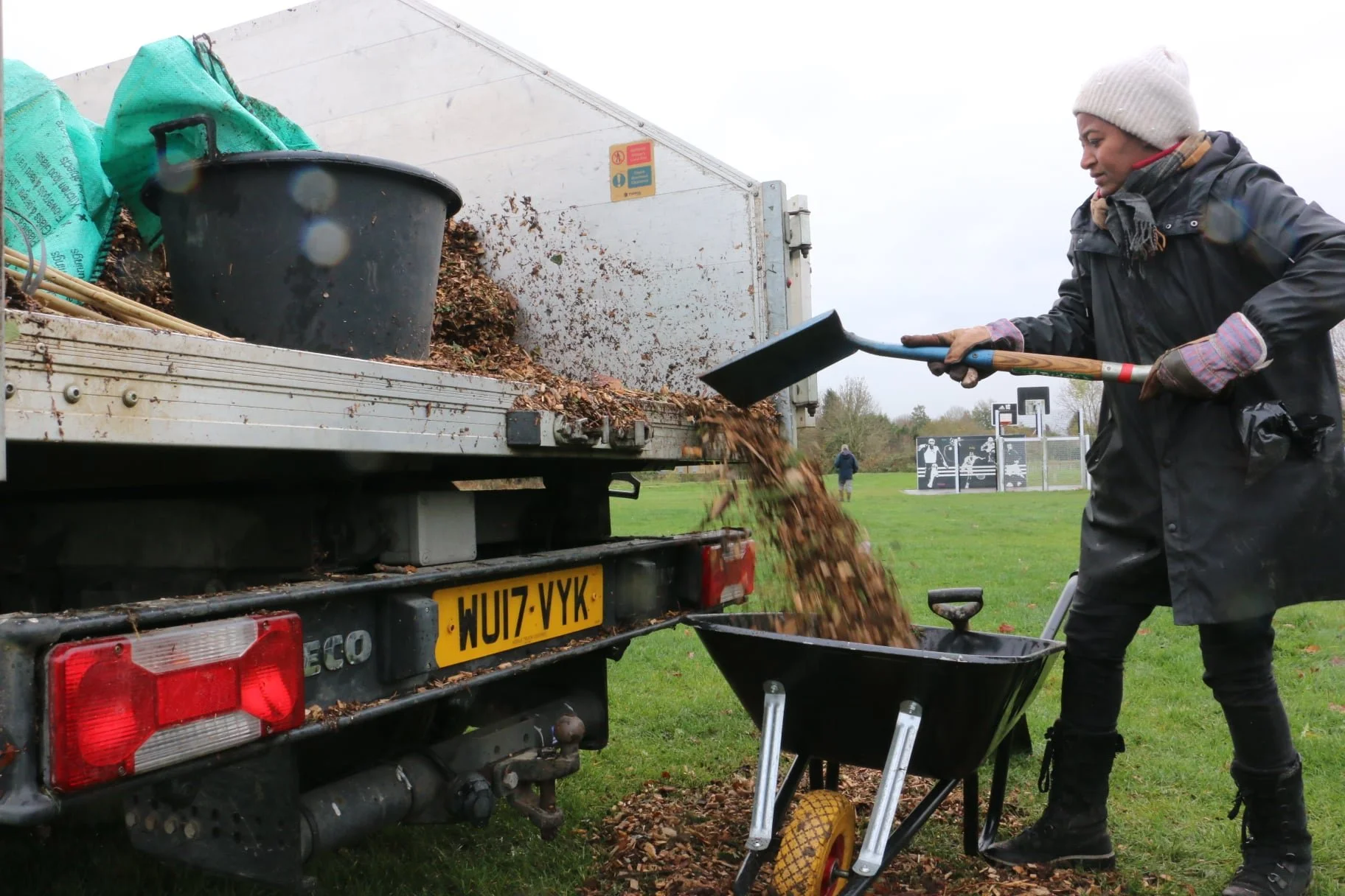 Person shoveling leaves and debris from a truck onto a wheelbarrow in a park.