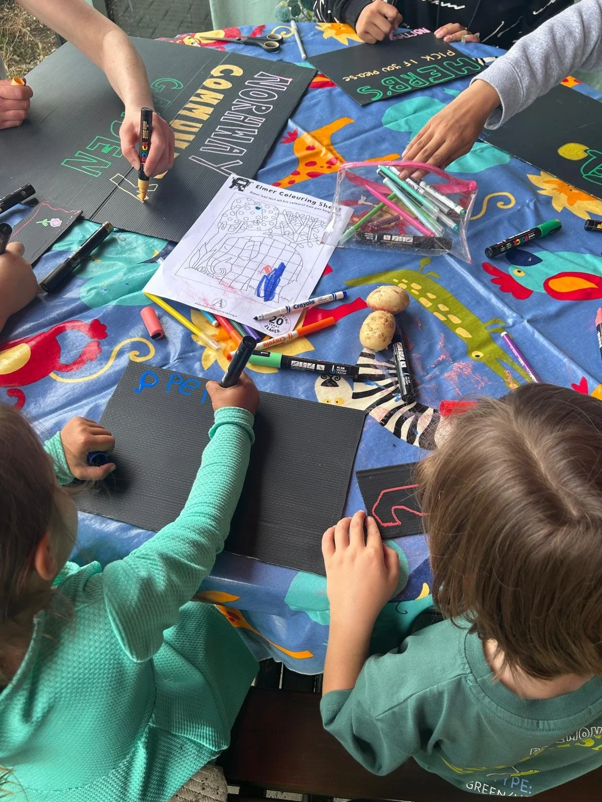 Children drawing and coloring on blackboards at a table covered with a colorful tablecloth with animal designs, with markers, scissors, and potatoes scattered across the table.