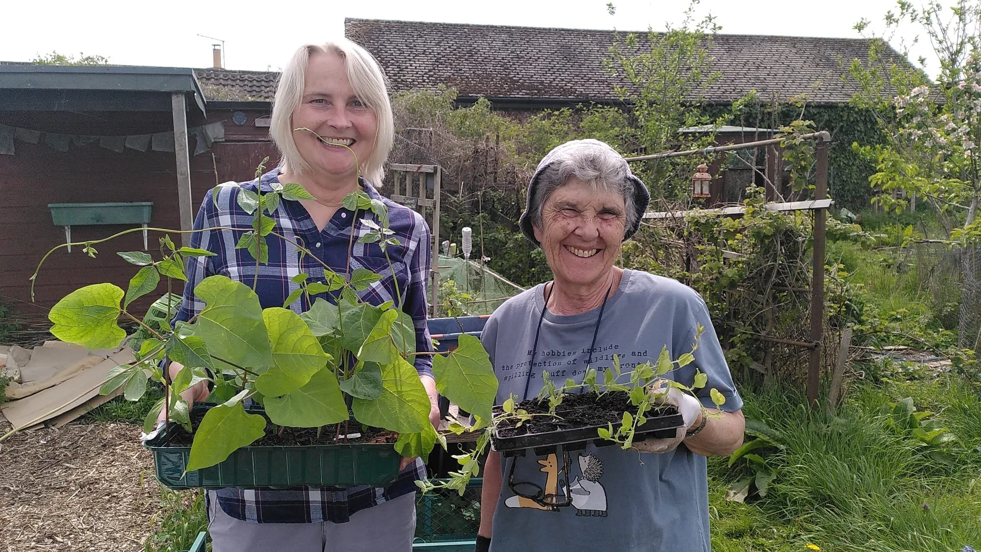 Two women smiling and holding trays with plants in a backyard garden.