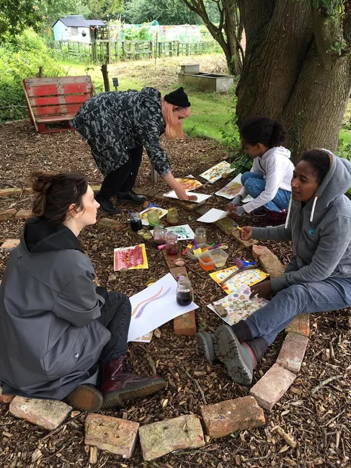 Four people sitting and standing around a small fire pit, engaged in outdoor craft activities, with colorful paints, brushes, and craft paper, under a large tree in a garden setting.