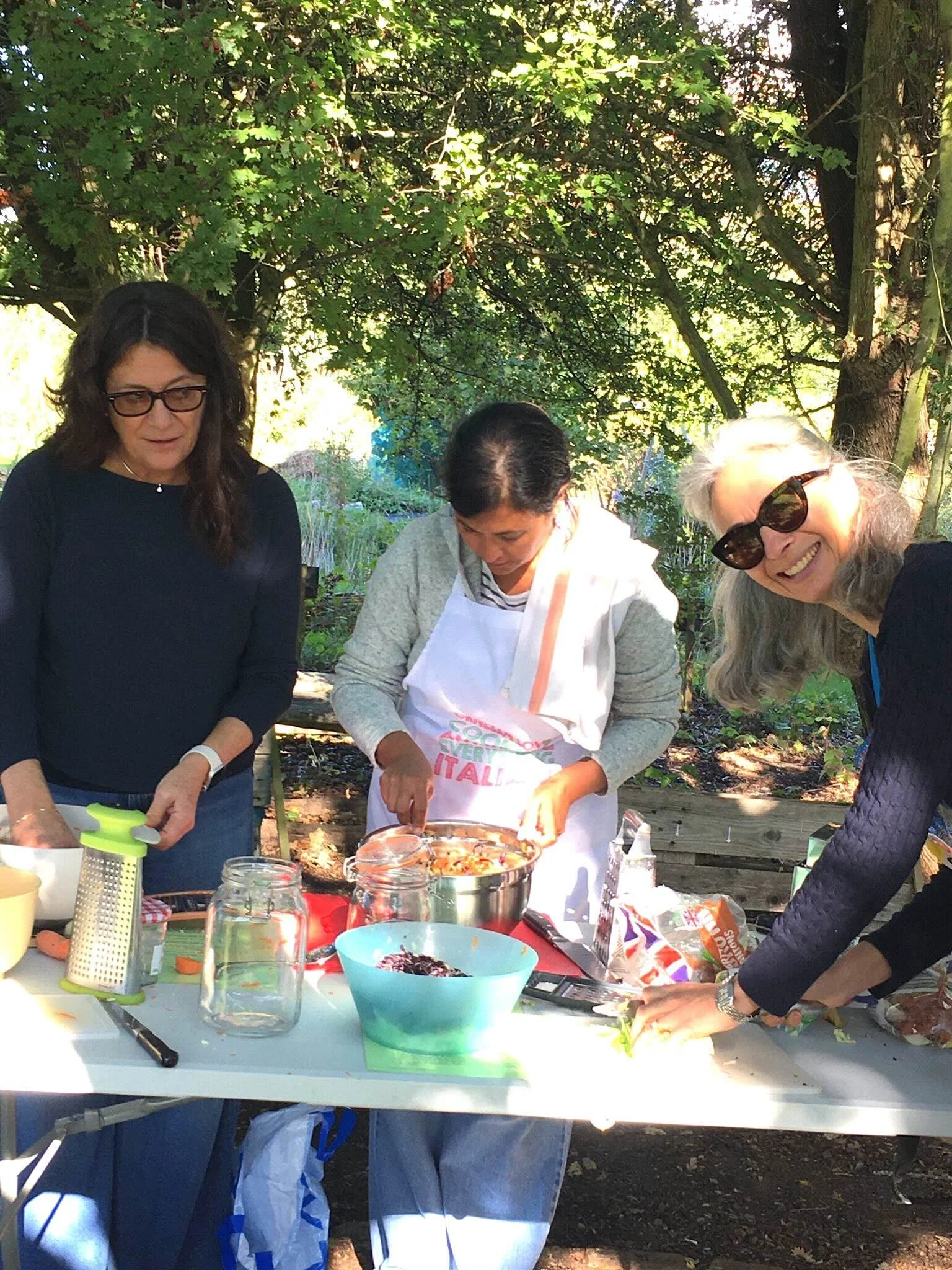 Three women are preparing food outdoors at a table. They are surrounded by trees and sunlight. One woman is smiling, another is focused on cooking, and the third woman is looking down at the table.
