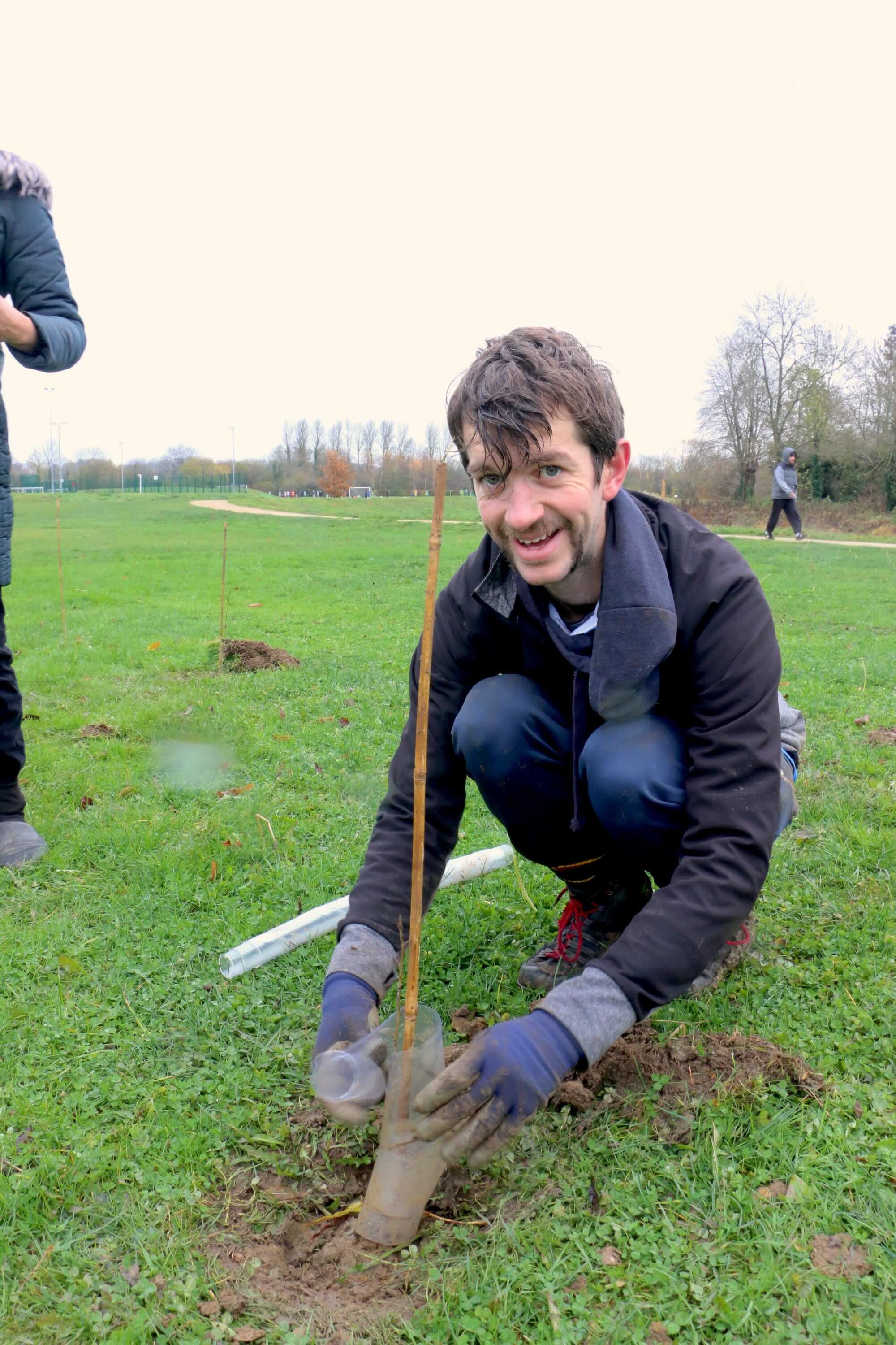 A man with brown hair and a beard planting a young tree in the ground, smiling and wearing a black jacket and gloves, in a park with green grass and other trees and people in the background.