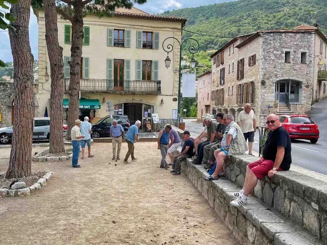 Group of men playing pétanque on a small dirt court in a European village square, with old buildings and parked cars in the background.