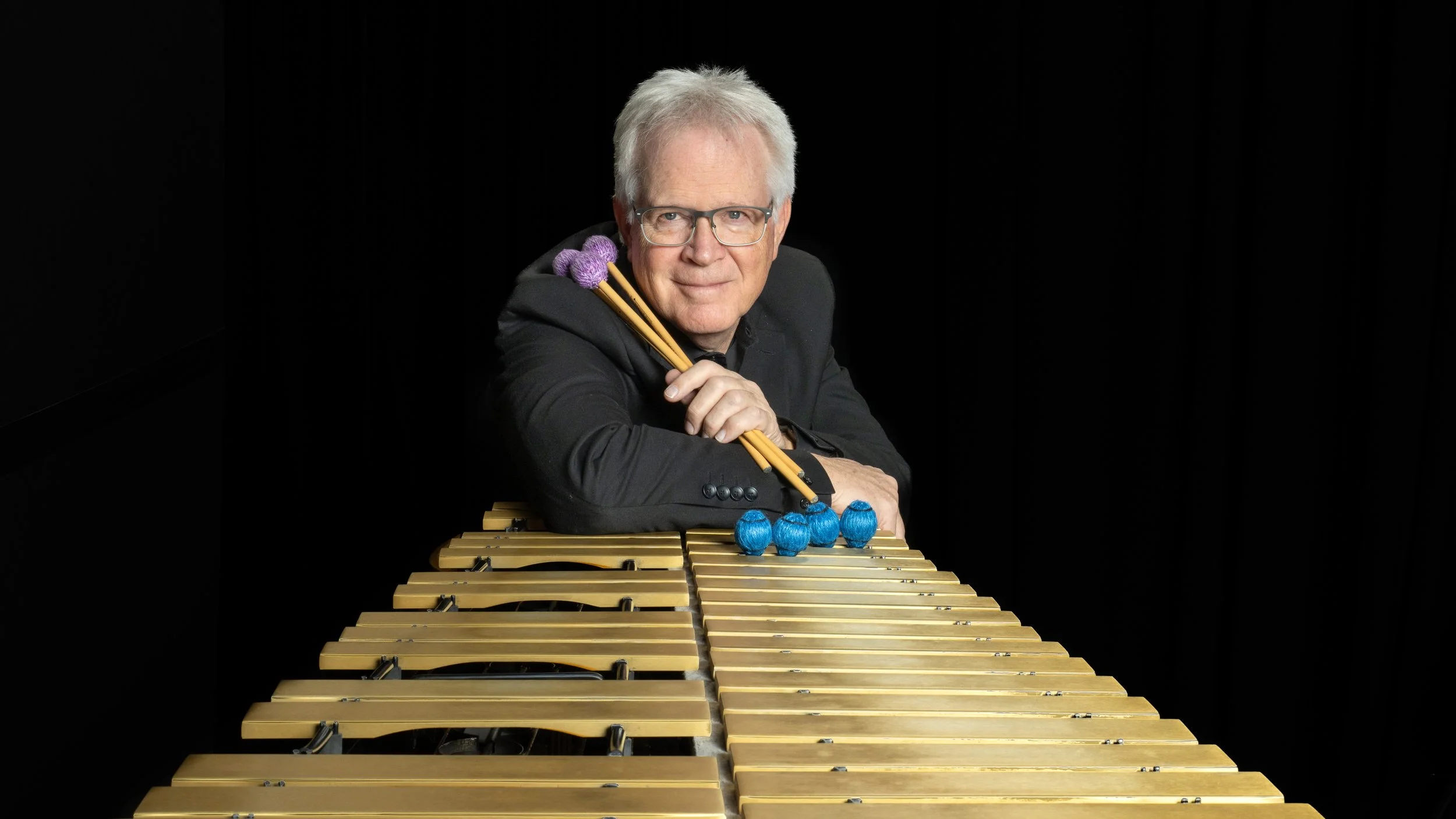 A man with glasses and white hair plays a vibraphone, holding purple and blue mallets, against a black background.