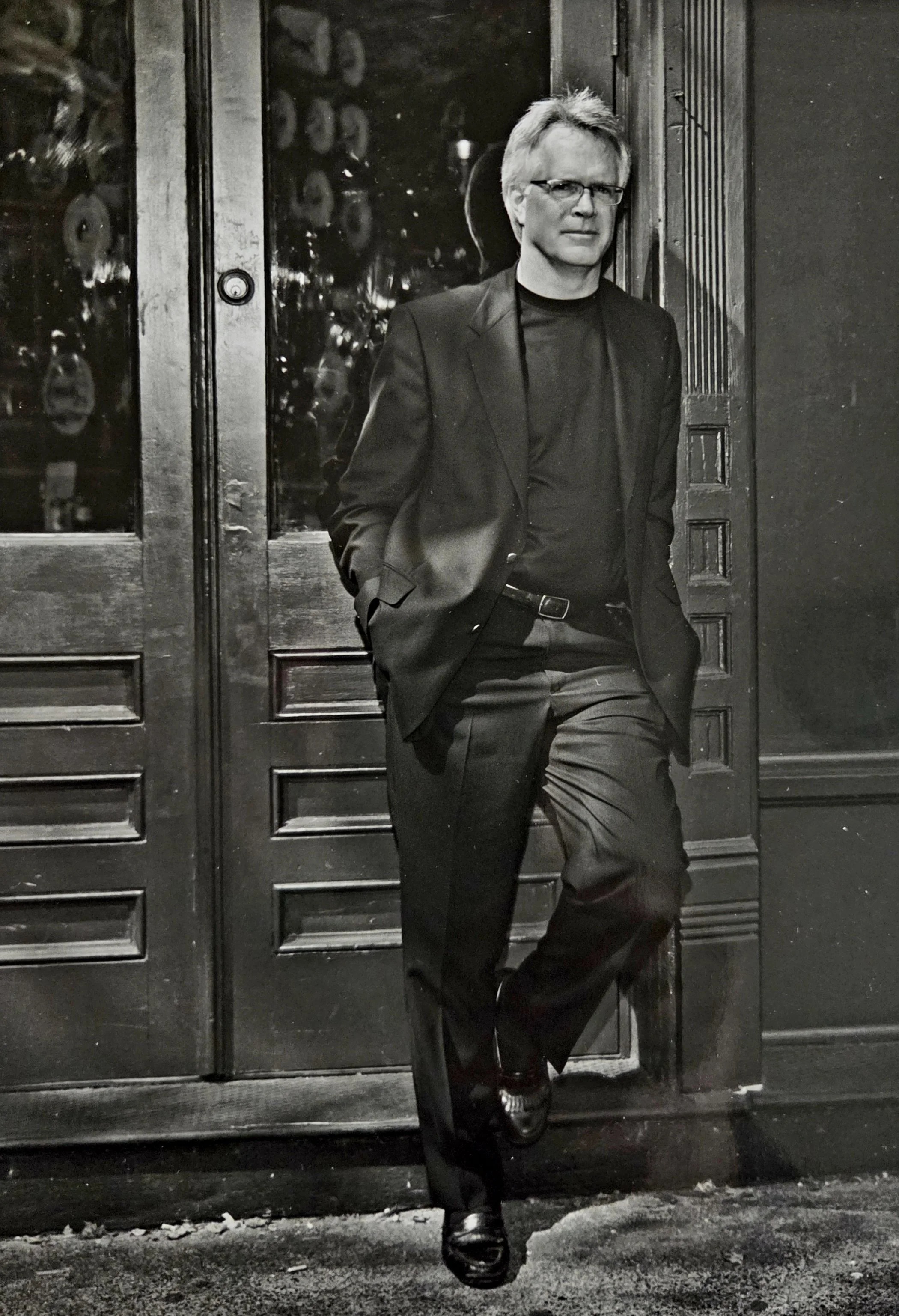 A man in a black suit and glasses leaning against a wooden door in black and white photo.