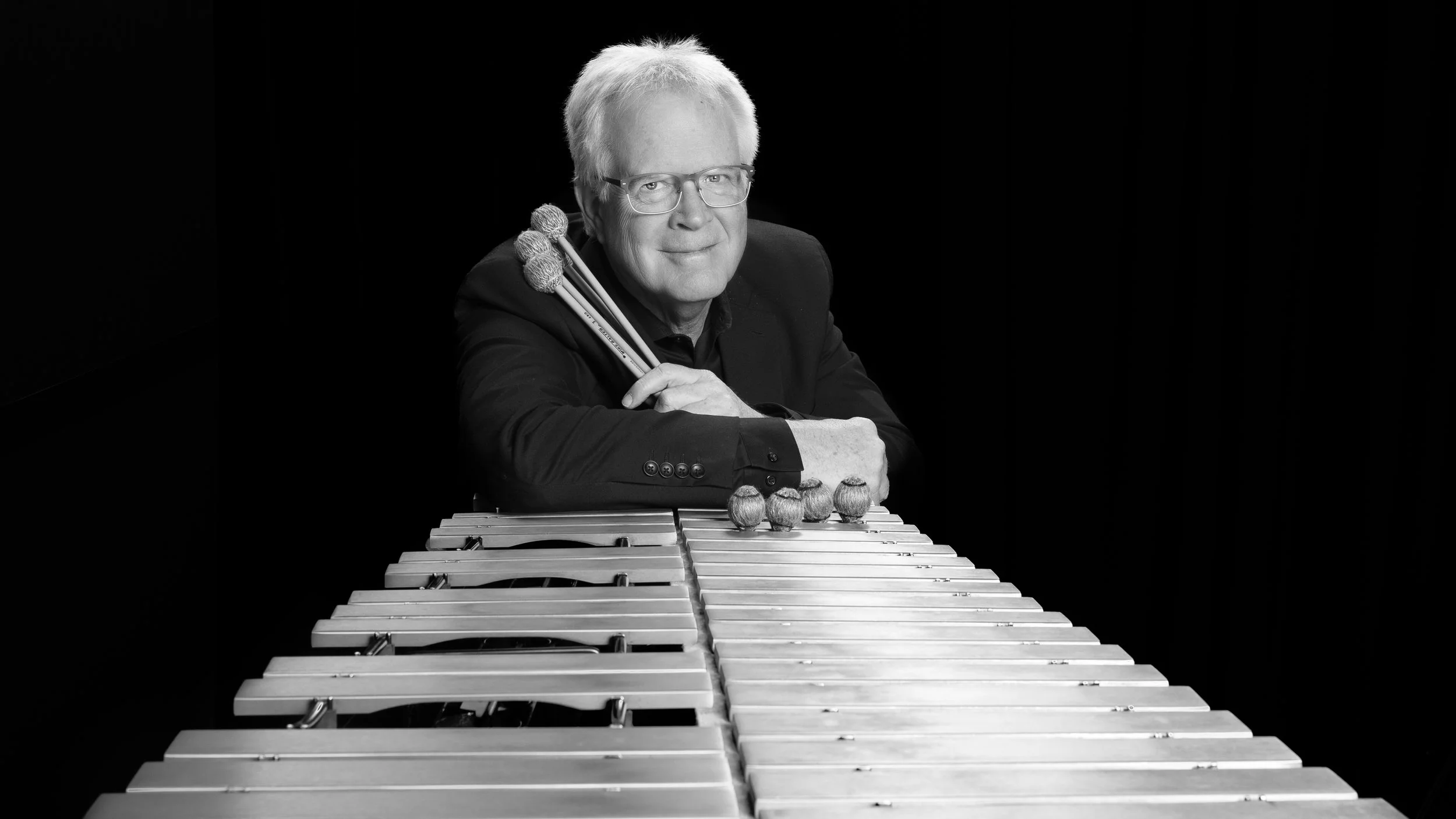 A black and white portrait of a man with white hair and glasses, sitting at a vibraphone with mallets resting on his shoulder, against a black background.