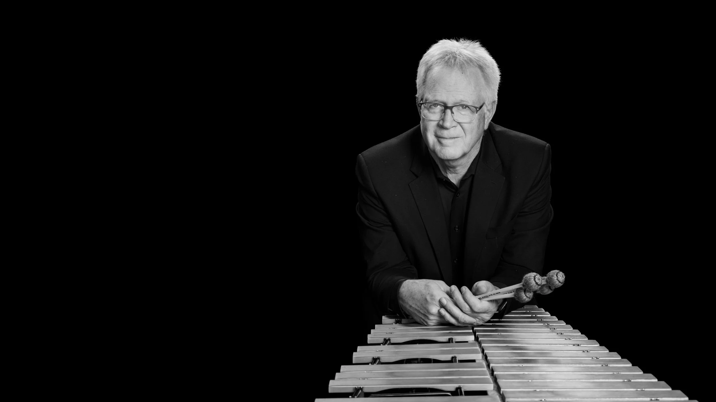 Black and white photo of a man playing a xylophone, holding mallets, against a black background.