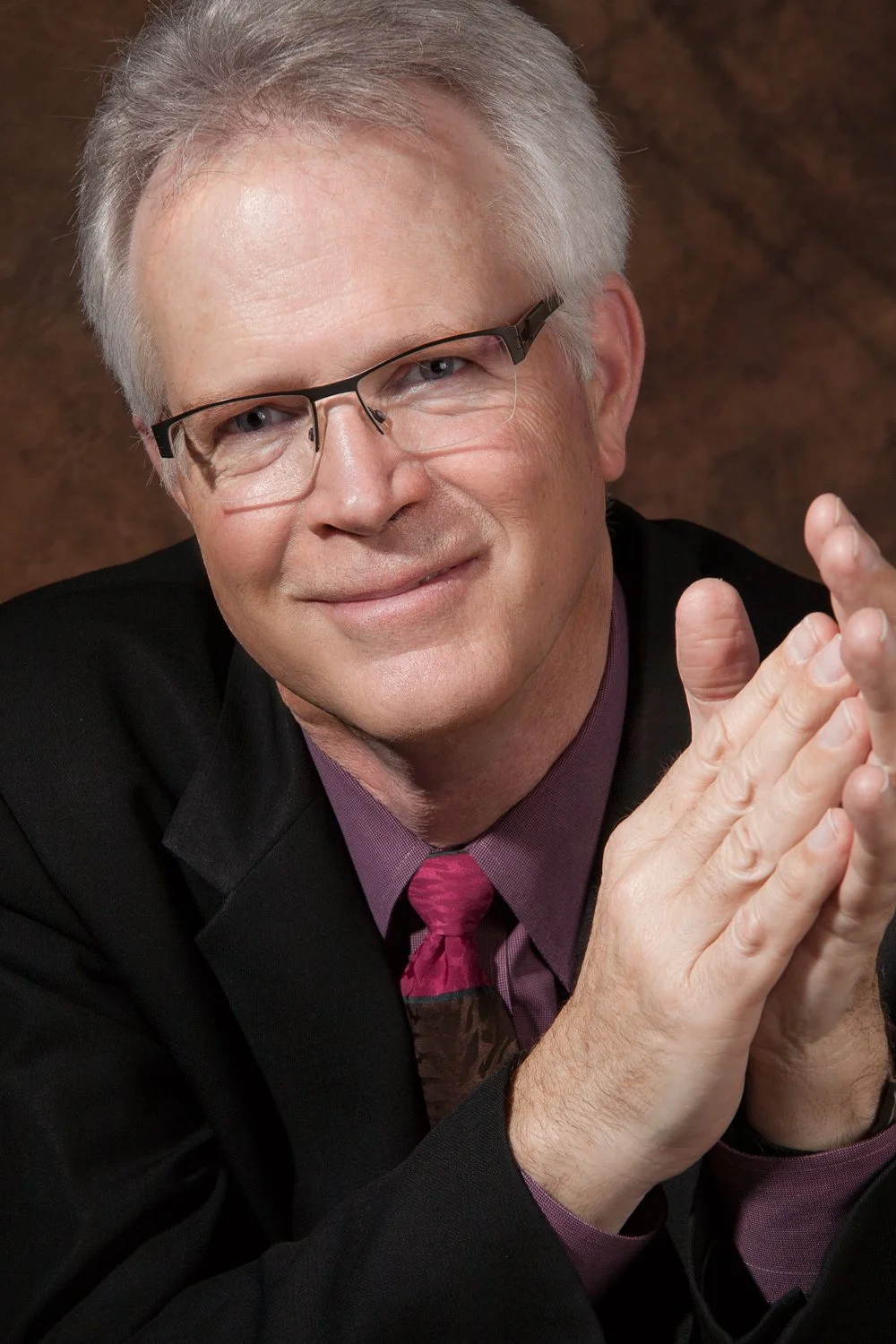A man with gray hair and glasses smiling, wearing a black suit, purple shirt, and red tie, with clasped hands.