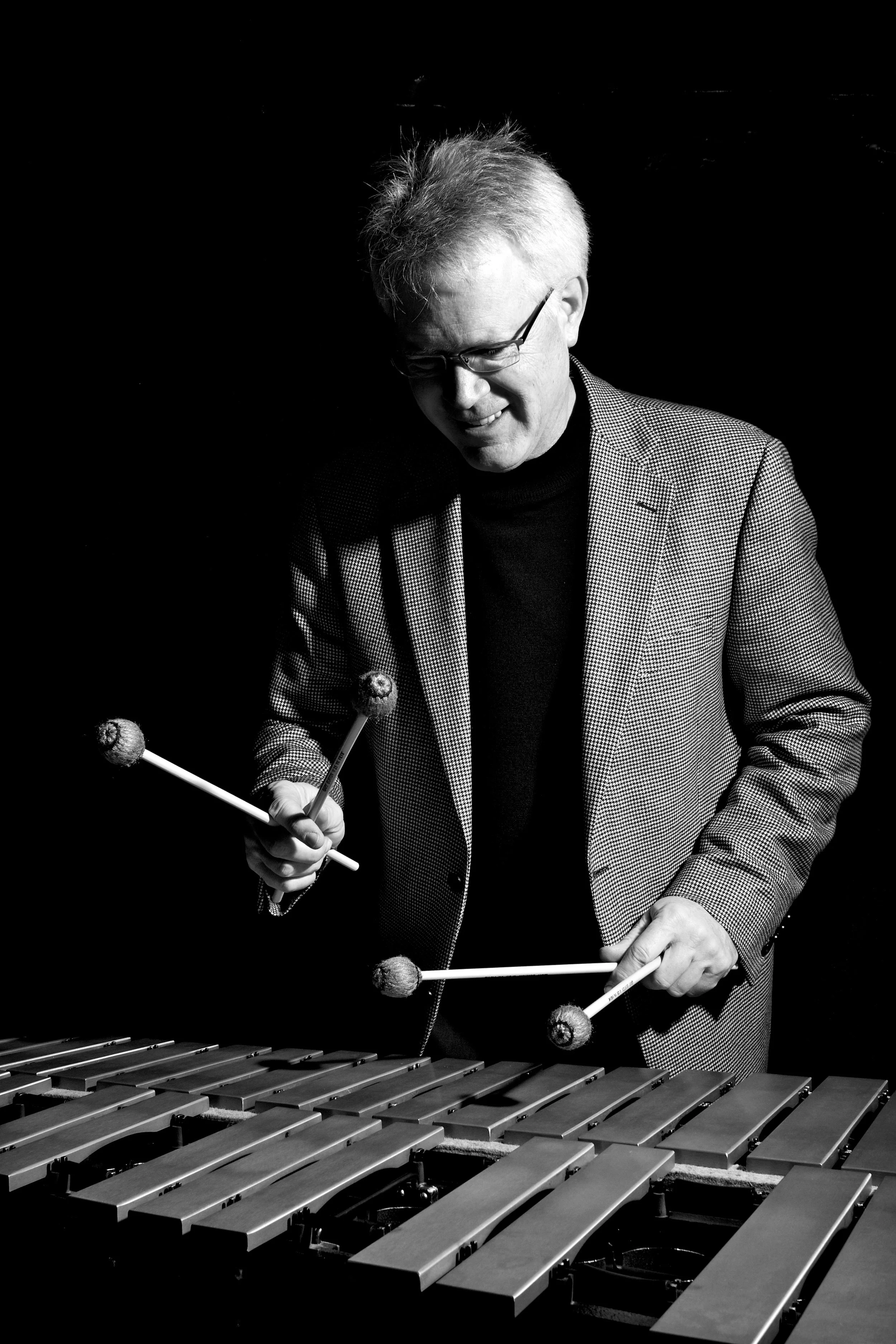 A man playing a vibraphone with mallets, wearing glasses, a checkered blazer, and a black turtleneck, smiling while focused on the instrument.