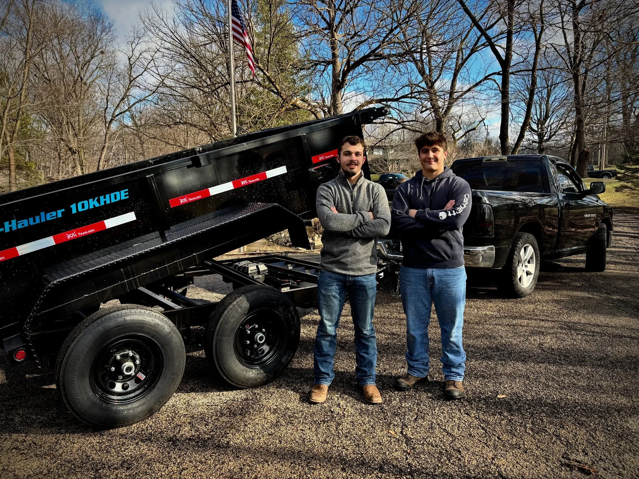 Two young men standing with arms crossed in front of a black pickup truck and a flatbed trailer on a gravel lot, with trees and an American flag in the background.