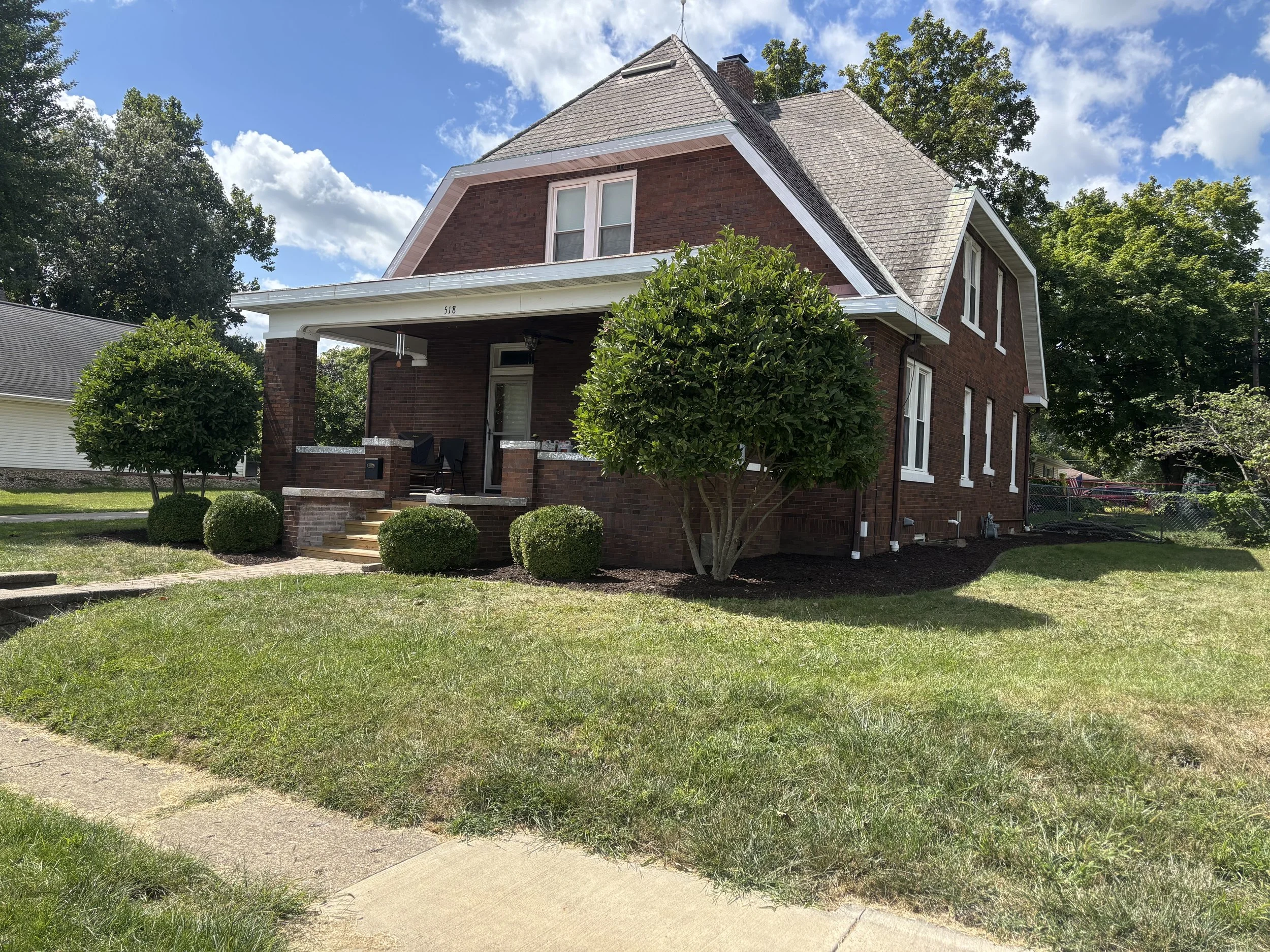 A two-story brick house with a front porch, surrounded by green bushes and lawn, under a partly cloudy blue sky.