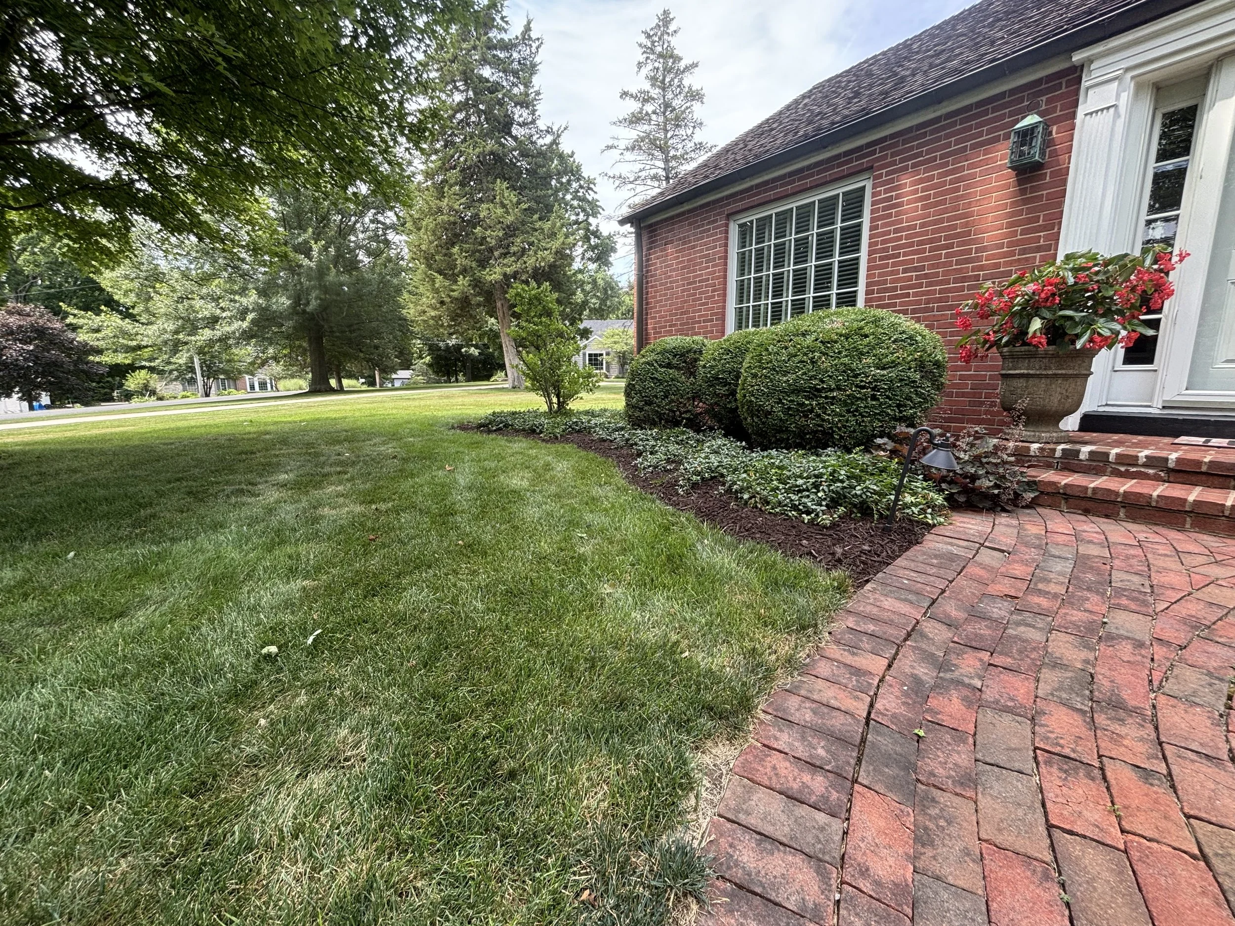 A neatly landscaped front yard with green grass, a curved brick walkway leading to a brick house, trimmed bushes, a flower pot with red flowers on the porch, and large trees in the background.