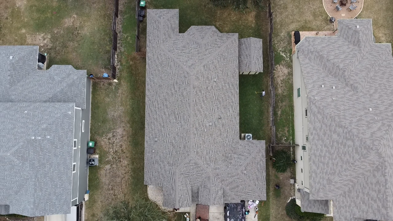 An aerial view of three suburban houses with gray shingle roofs, fenced backyards, and a lawn, with one house featuring an air conditioning unit and various backyard items visible.