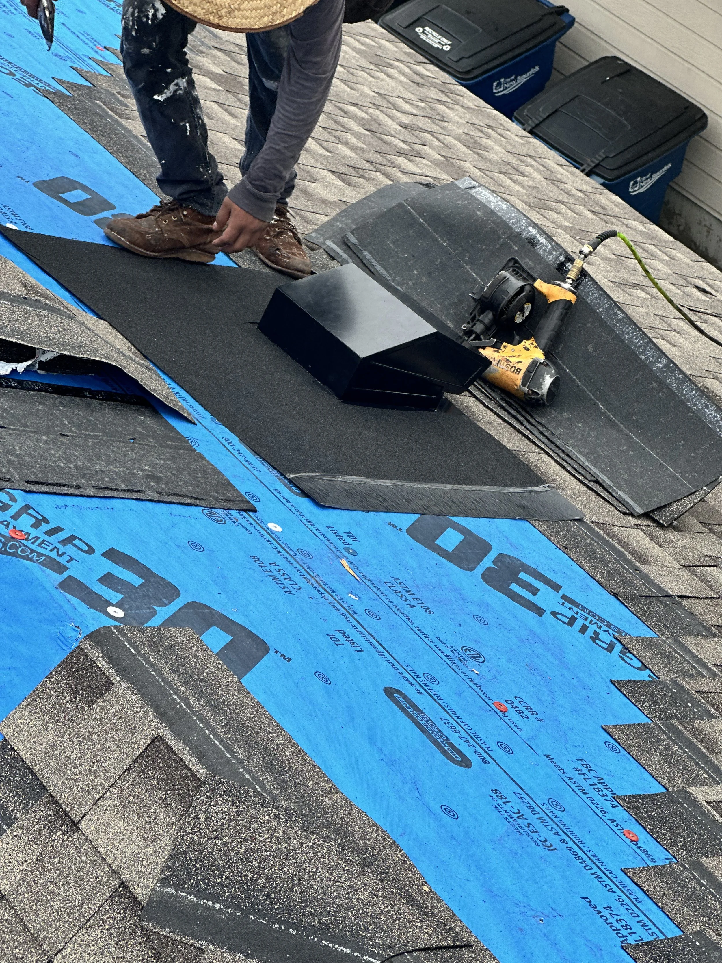 A worker installing asphalt roofing shingles on a roof, using a pneumatic nail gun and laying down underlayment.