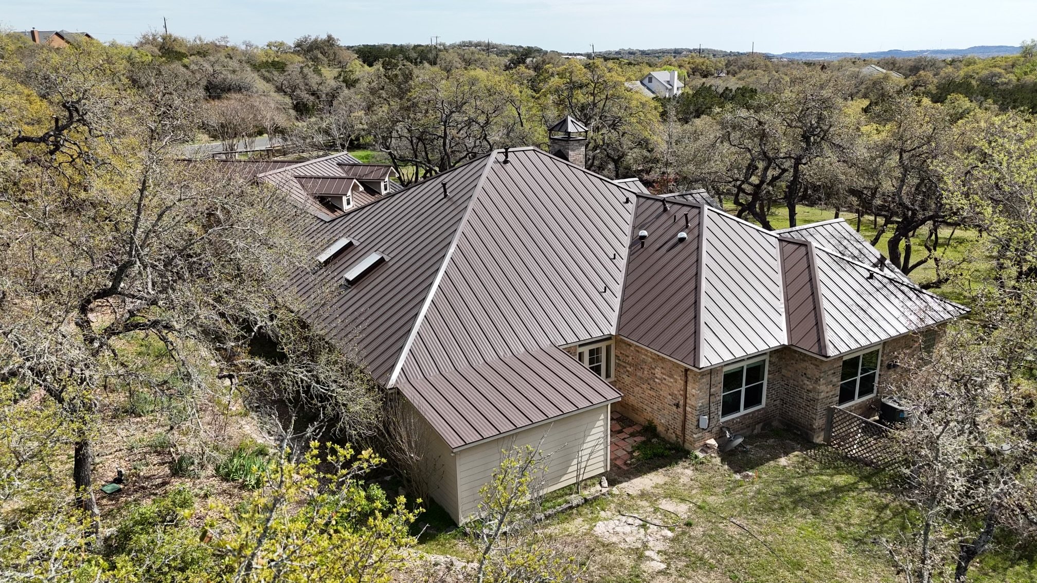 Aerial view of a house with a metal roof surrounded by trees and greenery in a rural area.