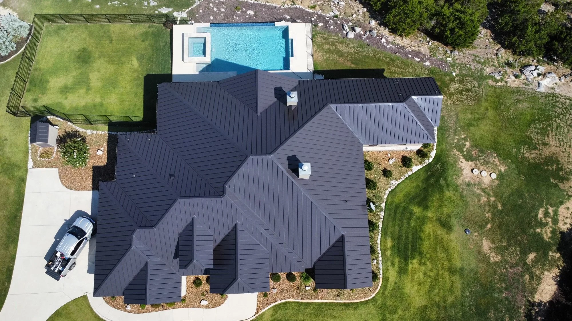 Aerial view of a house with a dark gray metal roof, a concrete driveway with a silver pickup truck, a fenced backyard with a swimming pool, a well-maintained lawn, and landscaping with trees and rocks.
