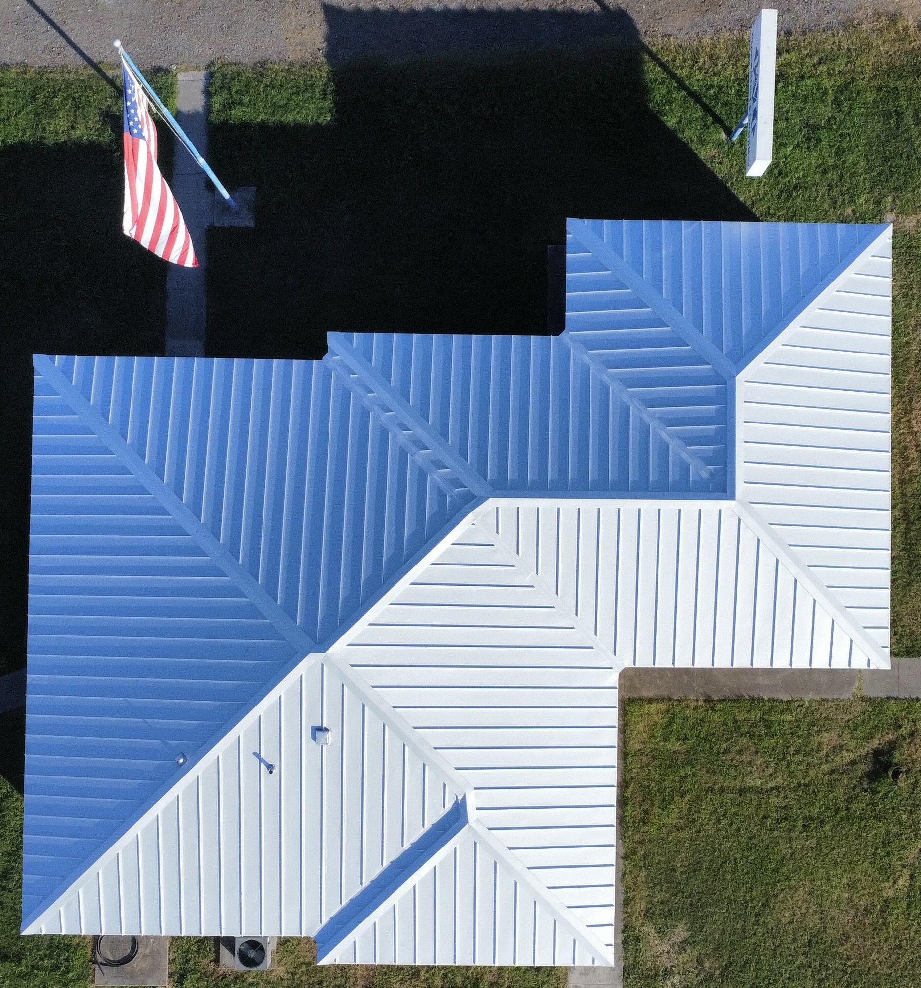 Top-down view of a house with white metal roof sections, beside a lawn, with an American flag on a pole.
