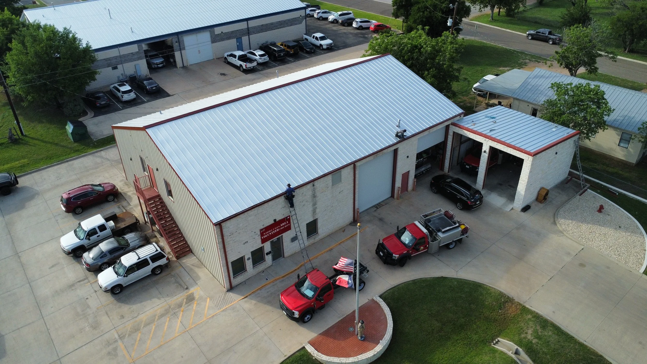 Aerial view of a commercial building with a metal roof, surrounded by parking lots and green lawns, with several vehicles parked and two workers cleaning the exterior walls.