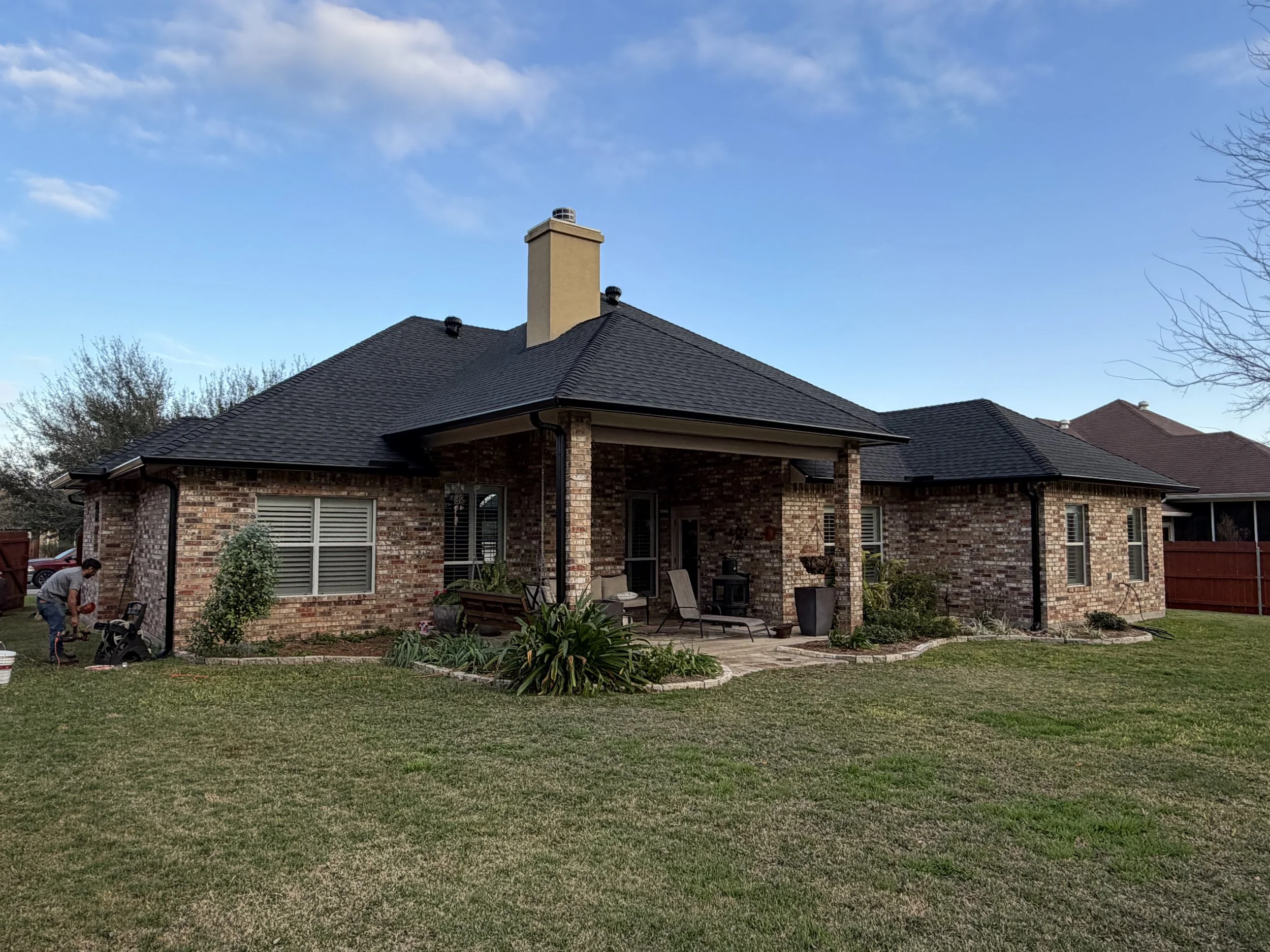 Brick house with black roof, backyard patio, lawn, and trees