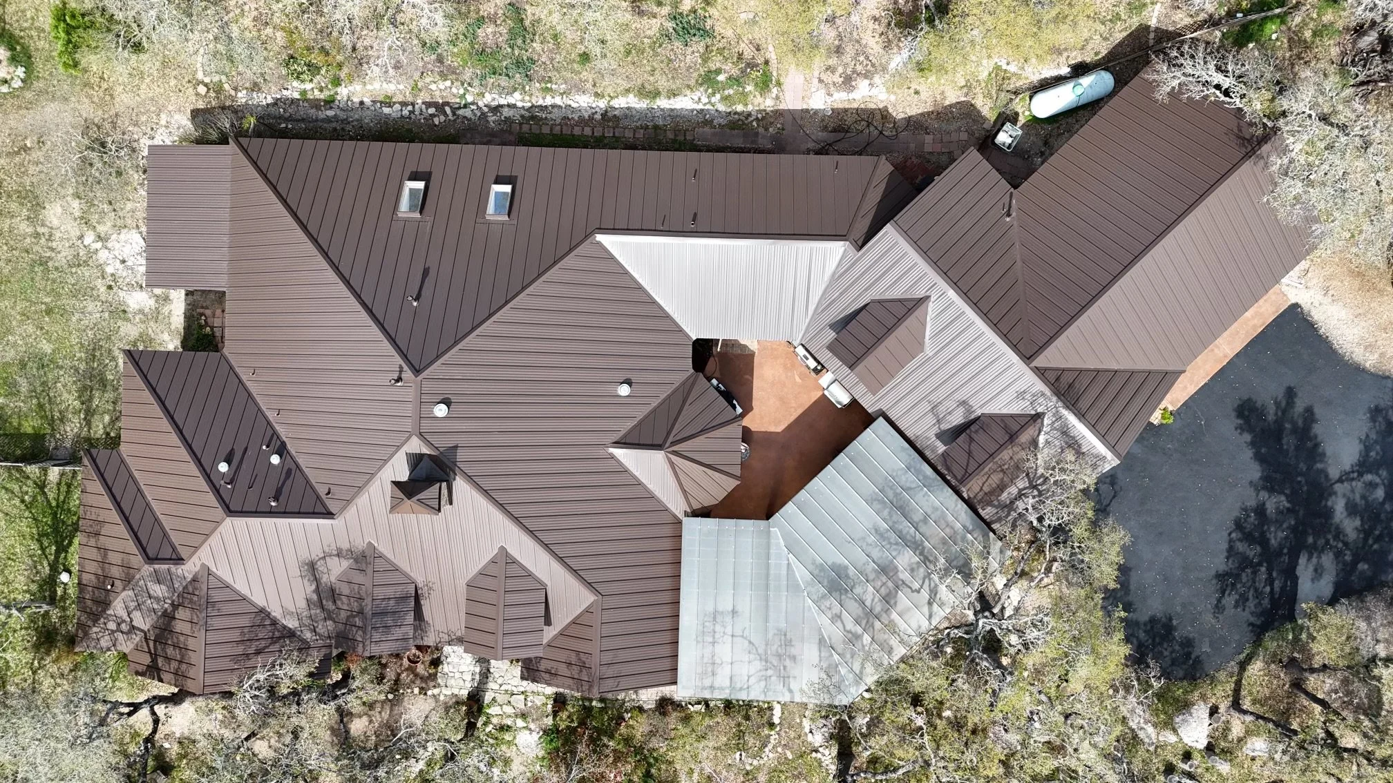 Aerial view of a house with a complex brown metal roof, multiple gables, and skylights, surrounded by trees, a driveway, and a small pond.