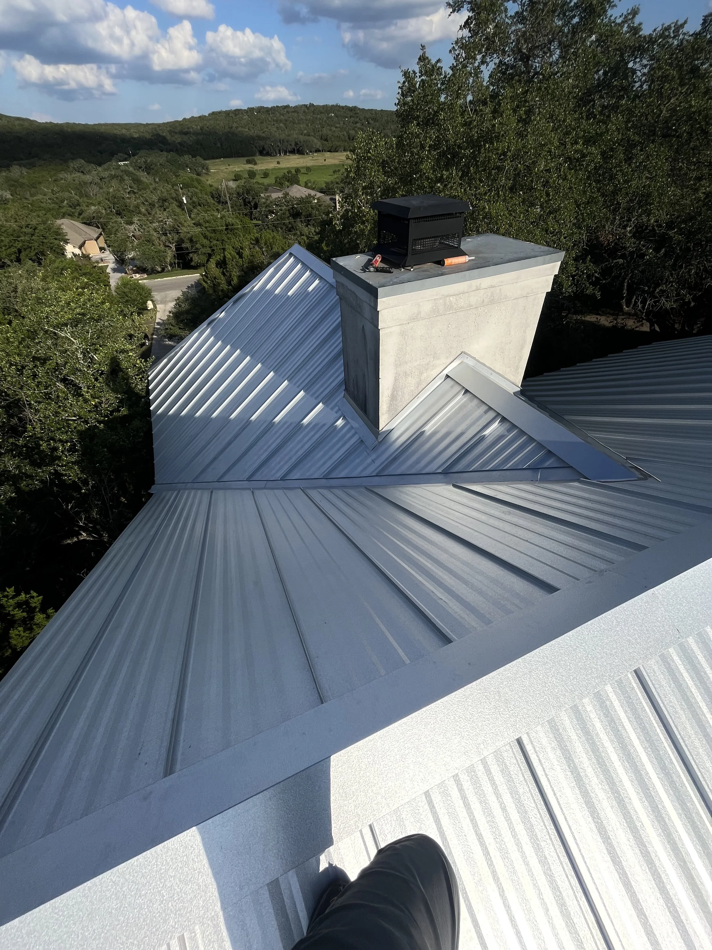 A metal rooftop with a chimney and a small ventilation unit, surrounded by trees and distant hills under a partly cloudy sky.