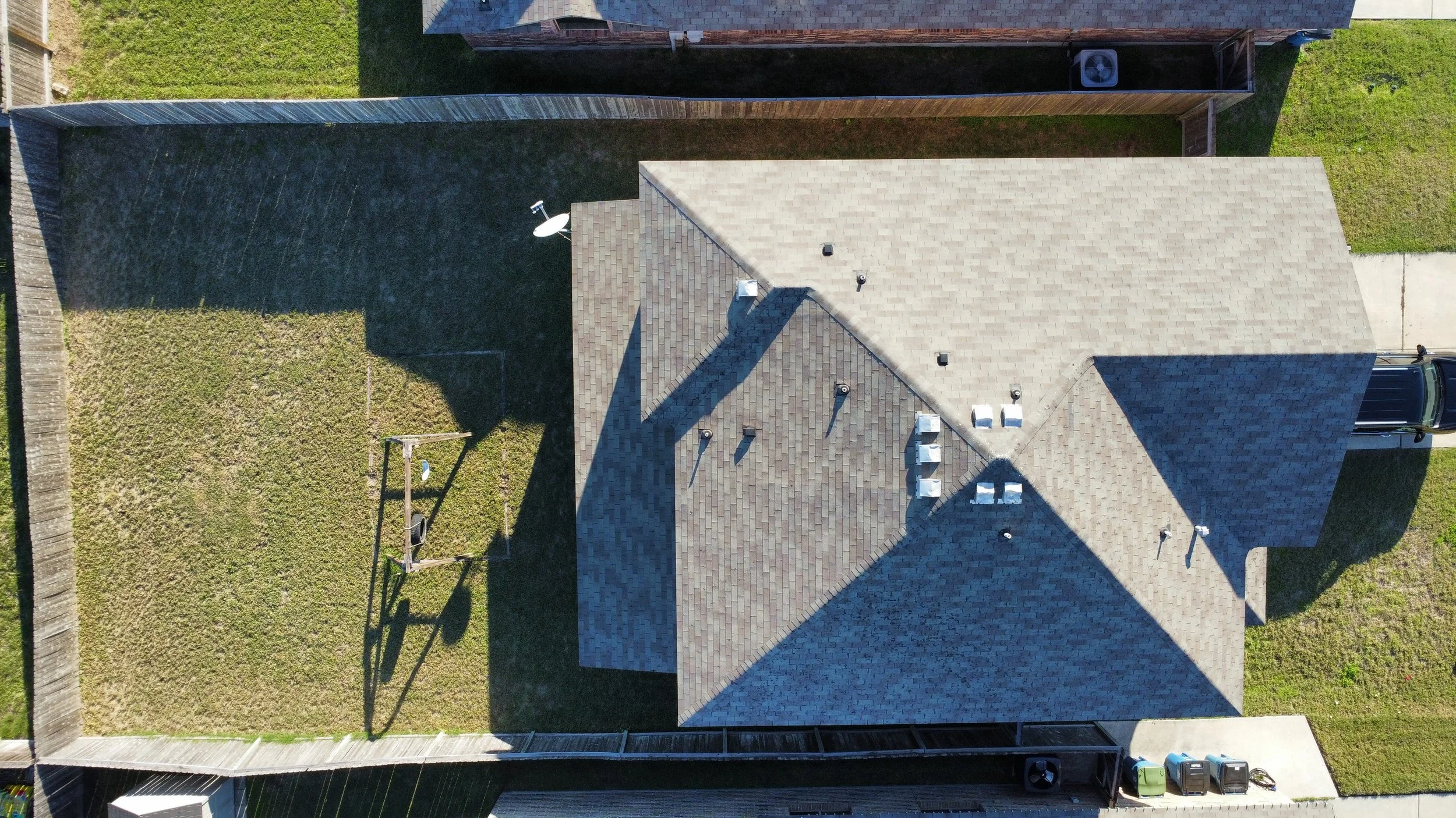 Top-down aerial view of a house with a grey shingled roof, backyard with grass, a swing set, and a satellite dish, surrounded by a wooden fence.