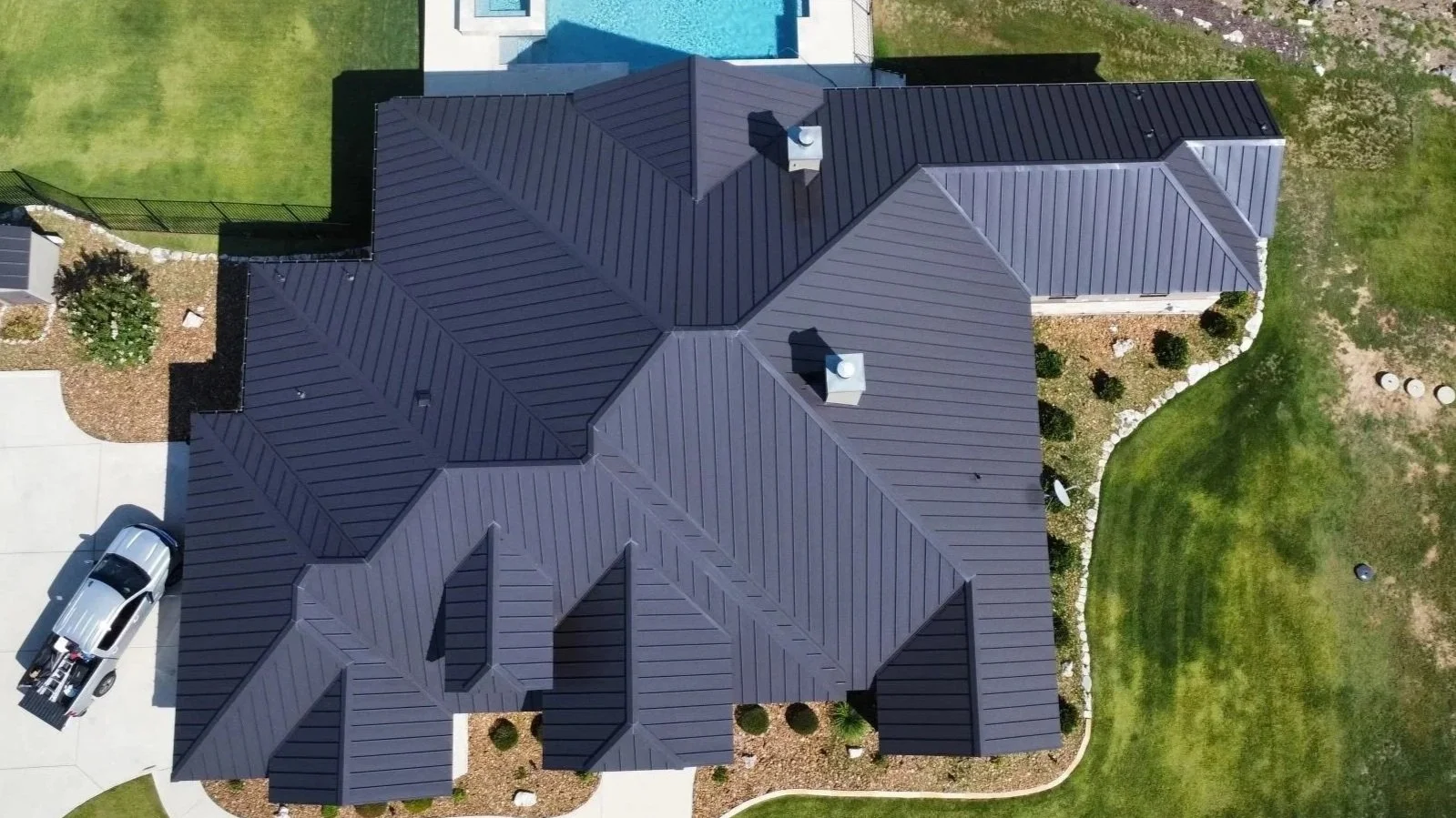 Aerial view of a house with a dark shingled roof, a driveway with a parked silver car, and surrounding green lawns and landscaped areas with bushes.