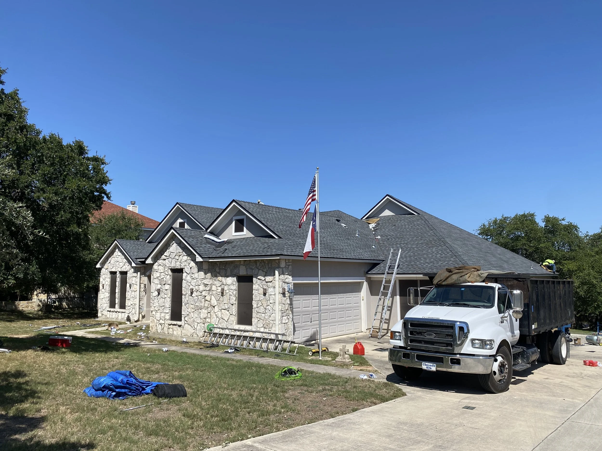 House under construction with workers, a truck, a ladder, and flags on a sunny day.