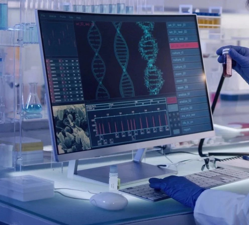Modern laboratory with a computer monitor displaying DNA sequences, graphs, and microscopic images. A scientist wearing gloves is holding a test tube.