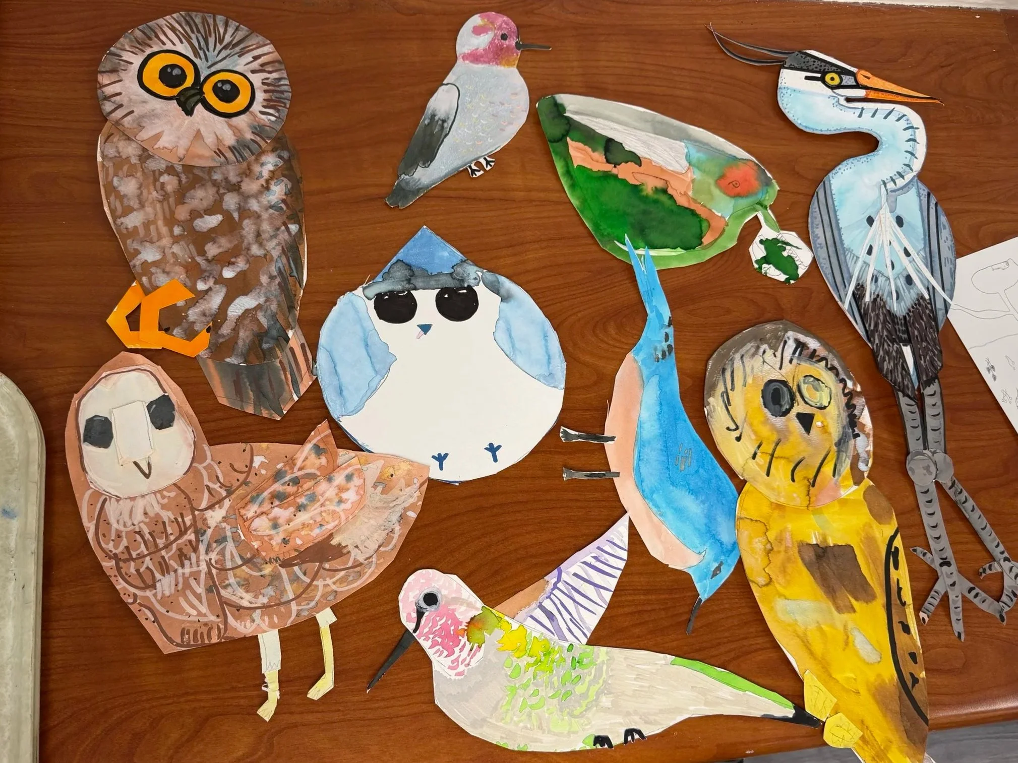 Colorful paper cutout artworks of various animals including an owl, a bird, a butterfly, a fox, a lion, and a pelican on a wooden surface.