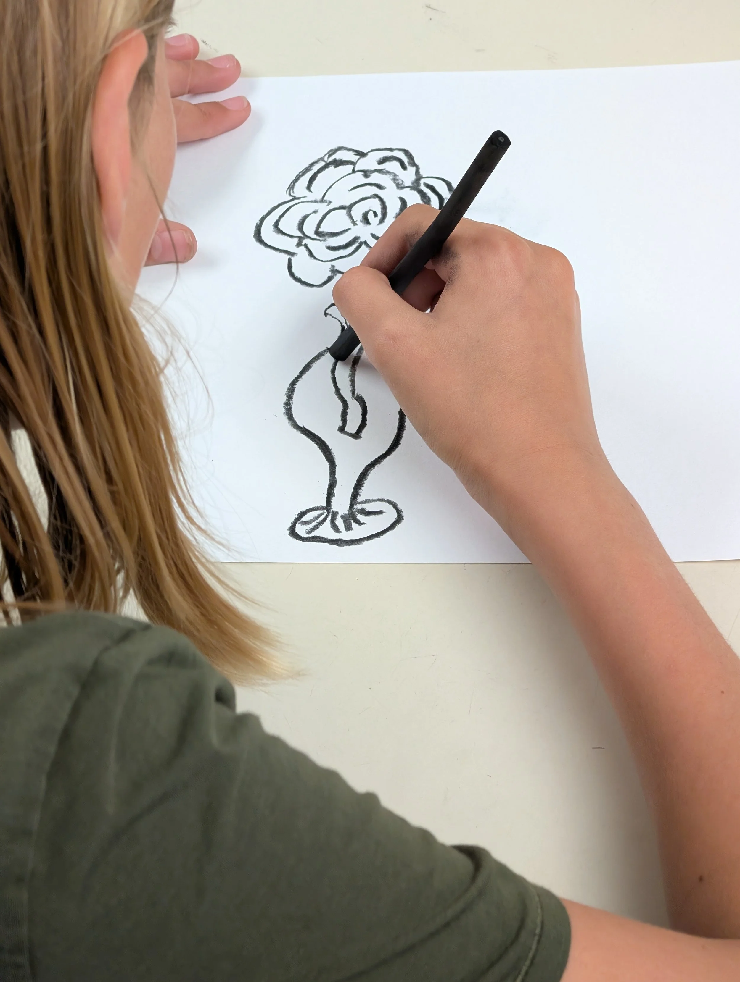 A person drawing a flower with a black marker on a white sheet of paper. The person has long blonde hair and is wearing a dark green shirt. The drawing depicts a large flower with multiple petals.