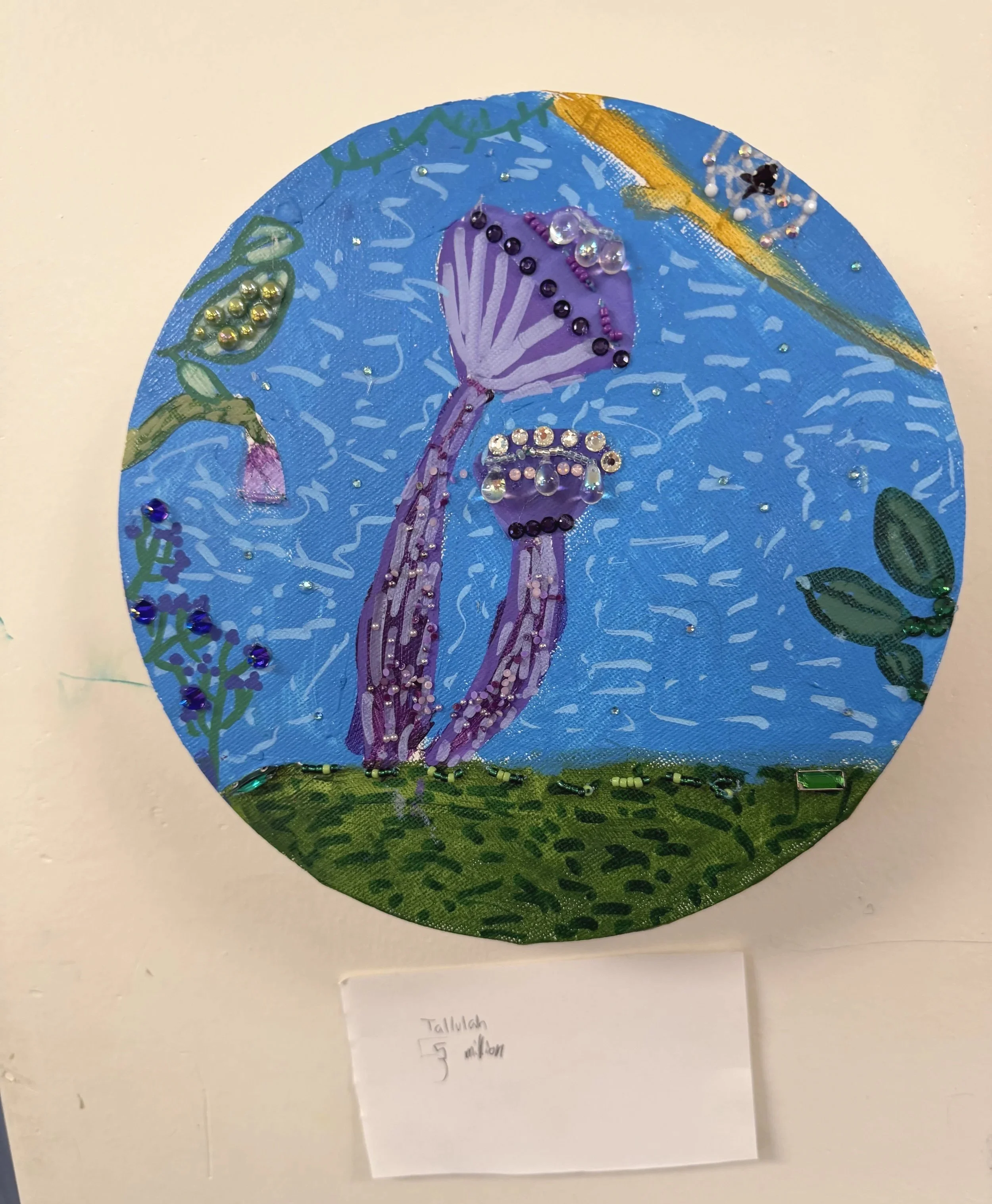 A circular art piece featuring a purple jellyfish with a string of black beads along its body and transparent beads simulating the jellyfish's bell, set against a blue background with green and yellow accents, decorated with various small rhinestones and sequins.