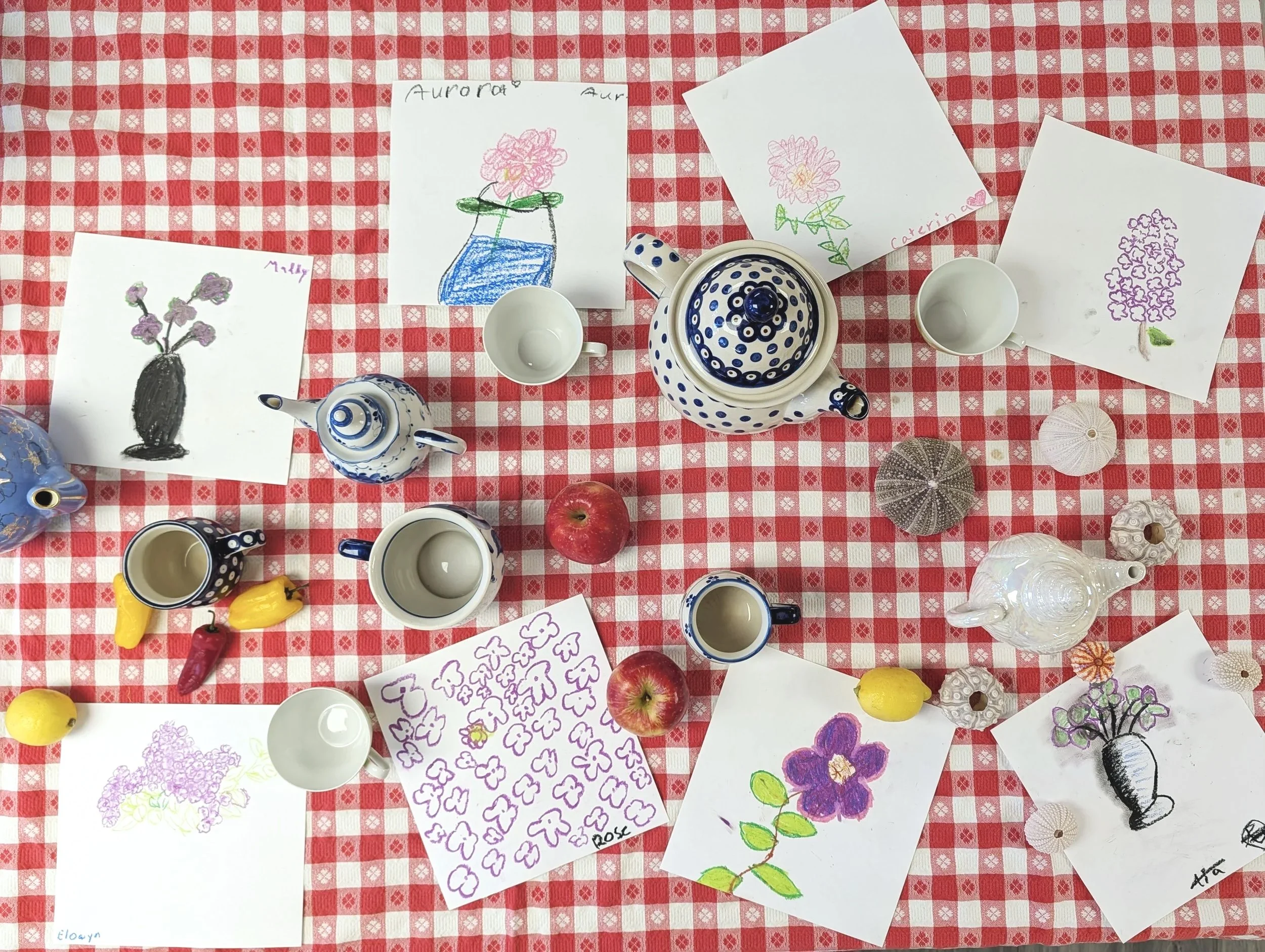 Colorful drawings of flowers, on white paper, placed on red and white checkered tablecloth, alongside teapots, cups, apples, yellow peppers, shells, a water bottle, and a red apple.