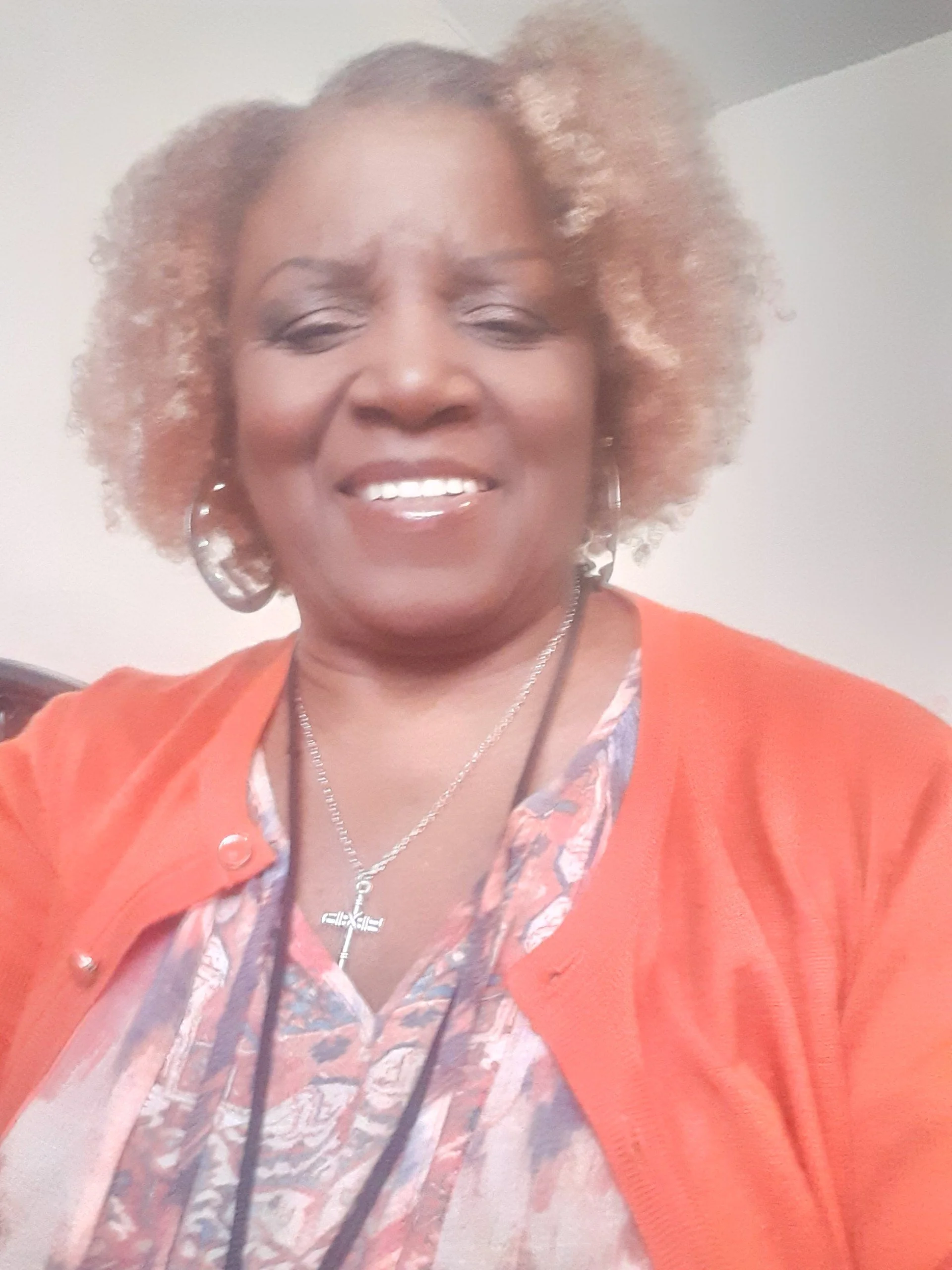 A smiling woman with curly, light brown hair with blonde highlights, wearing a patterned blouse, an orange cardigan, and silver jewelry, in front of a plain background.