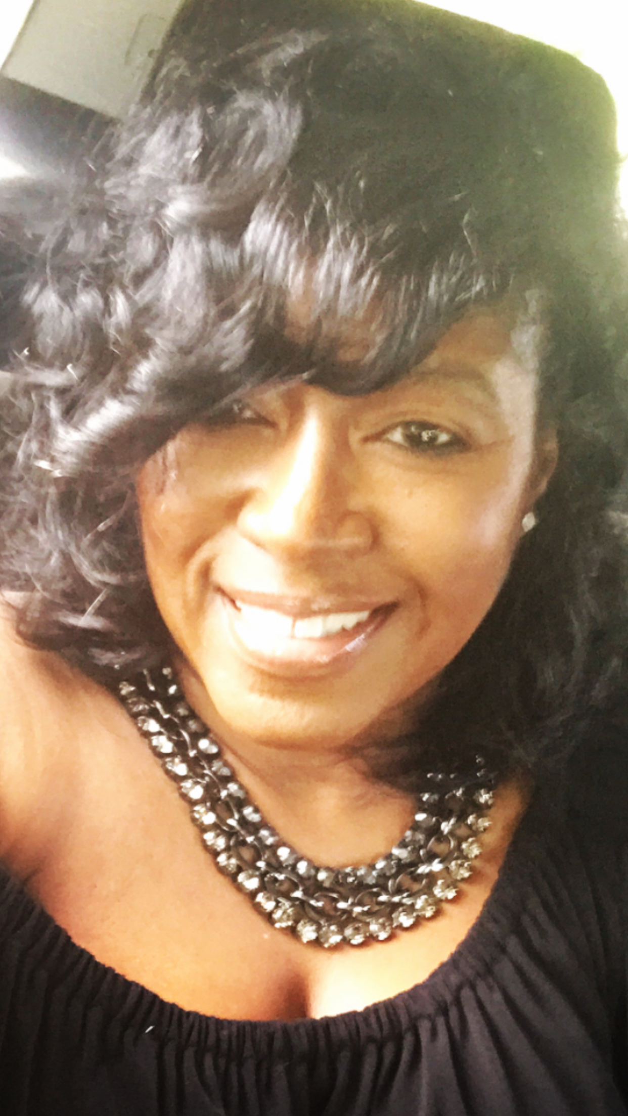 Close-up of a smiling woman with curly black hair, wearing a black top with a ruffled neckline and multiple layered chunky silver necklaces.