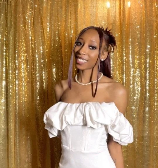 Young woman in a white off-the-shoulder dress posing in front of a glittery gold curtain.