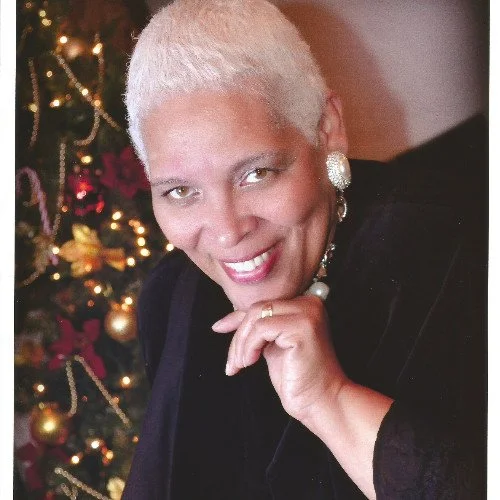 A woman with short white hair smiling, wearing large earrings and a black top, sitting in front of a decorated Christmas tree.