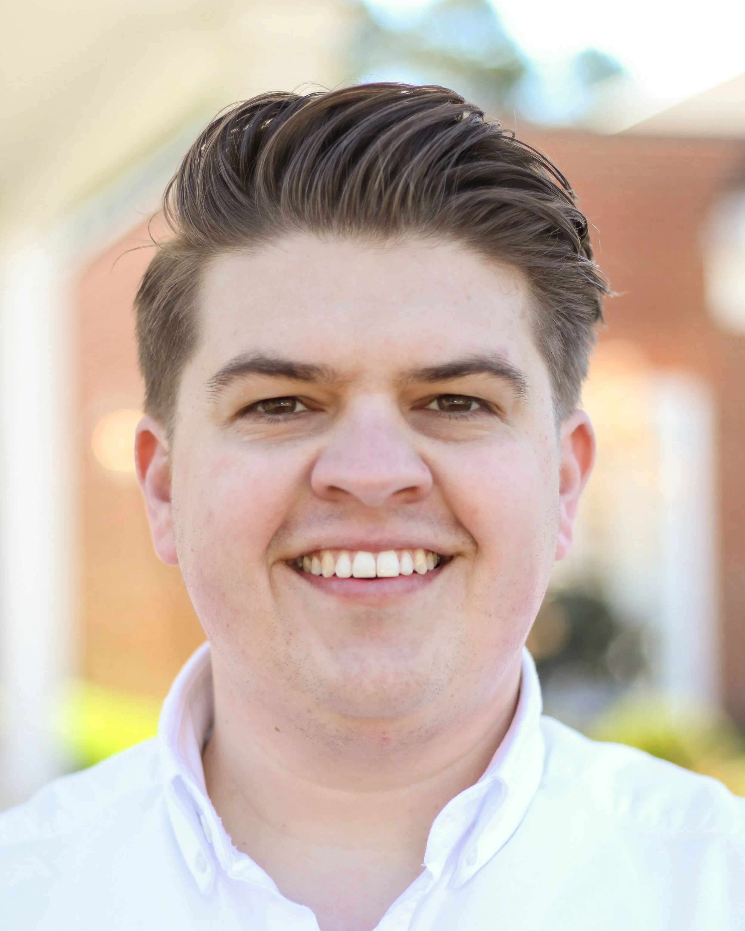 Close-up of a young man with styled brown hair, smiling, wearing a white collared shirt, outdoors with a blurred background of trees and building.