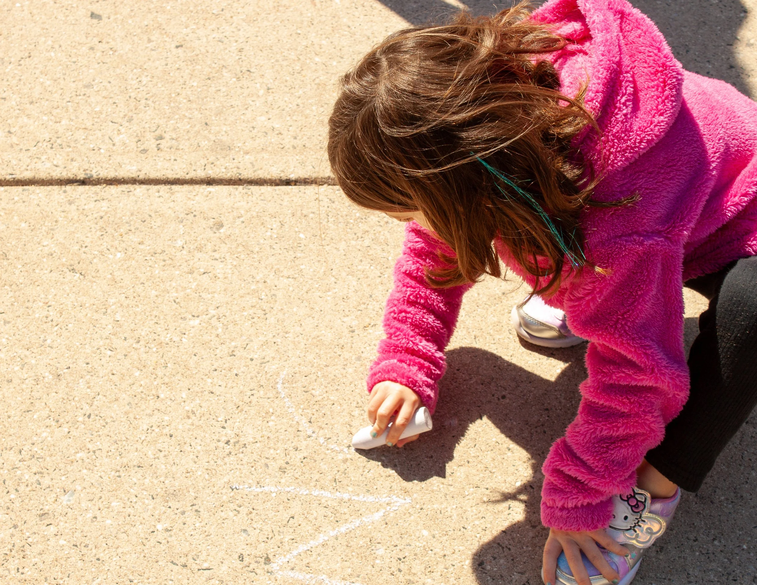 A young girl wearing a pink fuzzy jacket, lavender Hello Kitty shoes, and holding a bottle, crouched on a sidewalk.