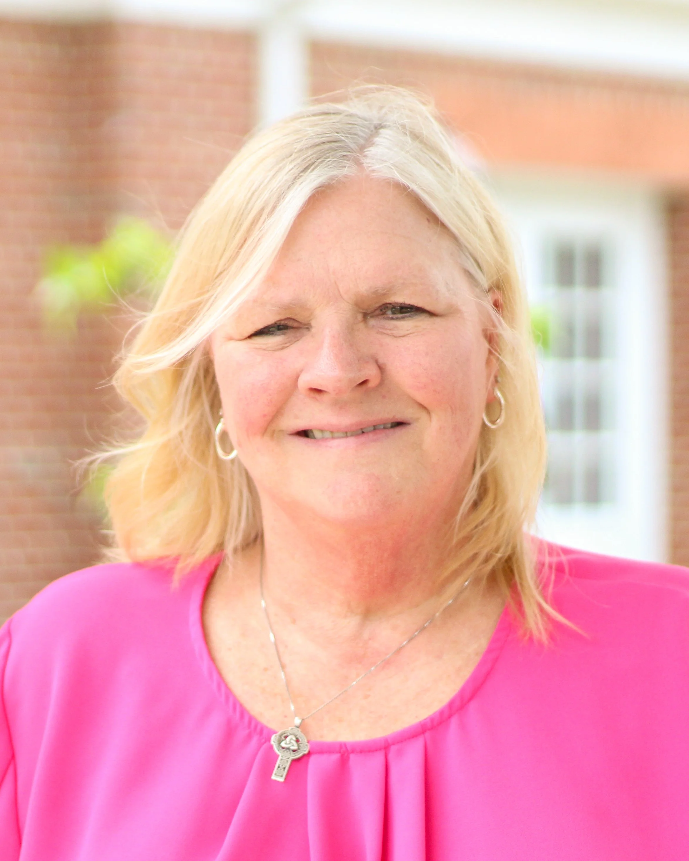 Close-up of a smiling woman wearing a pink top, necklace, and earrings, with blonde hair, outdoors with a brick building and window in the background.