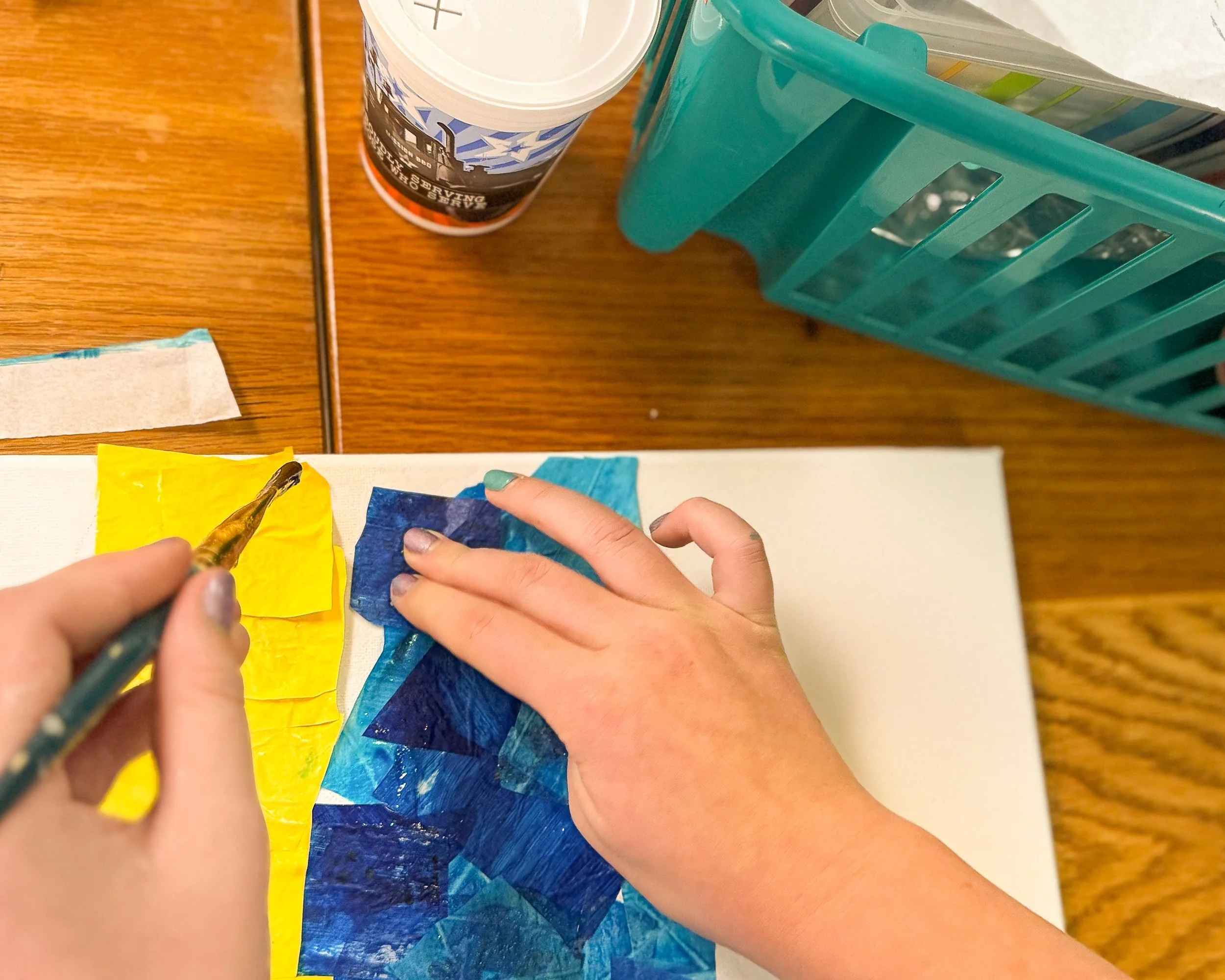 Person painting with blue and yellow paint on paper using a paintbrush. Hand seen with painted nails. Wooden surface, a cup, and a teal-colored plastic holder with art supplies in the background.